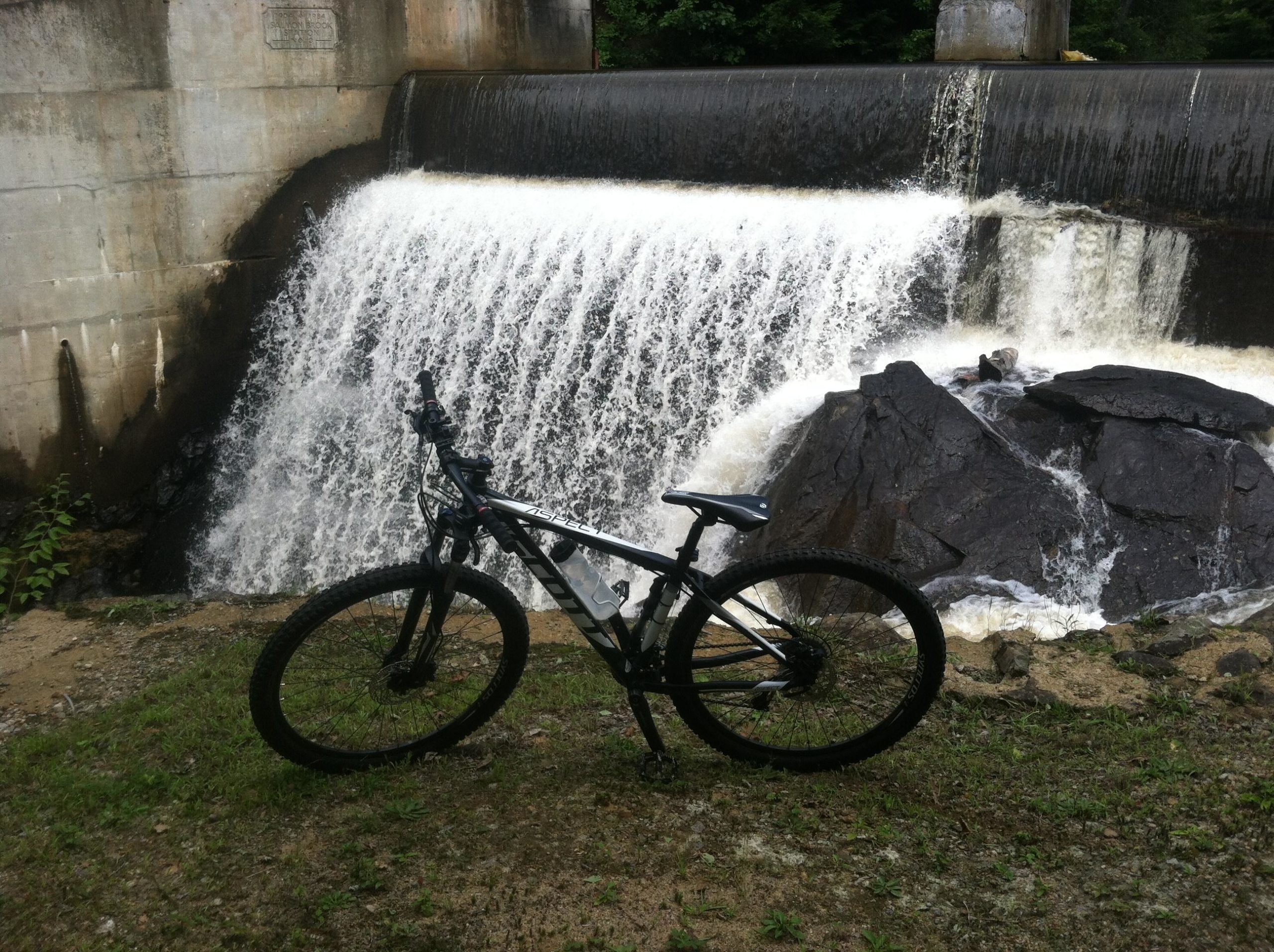 A black mountain bike parked on the grass near a waterfall, with water cascading over a concrete dam in the background. Rocks and greenery are visible beside the bike and the flowing water. Franklin Falls mountain bike trail.