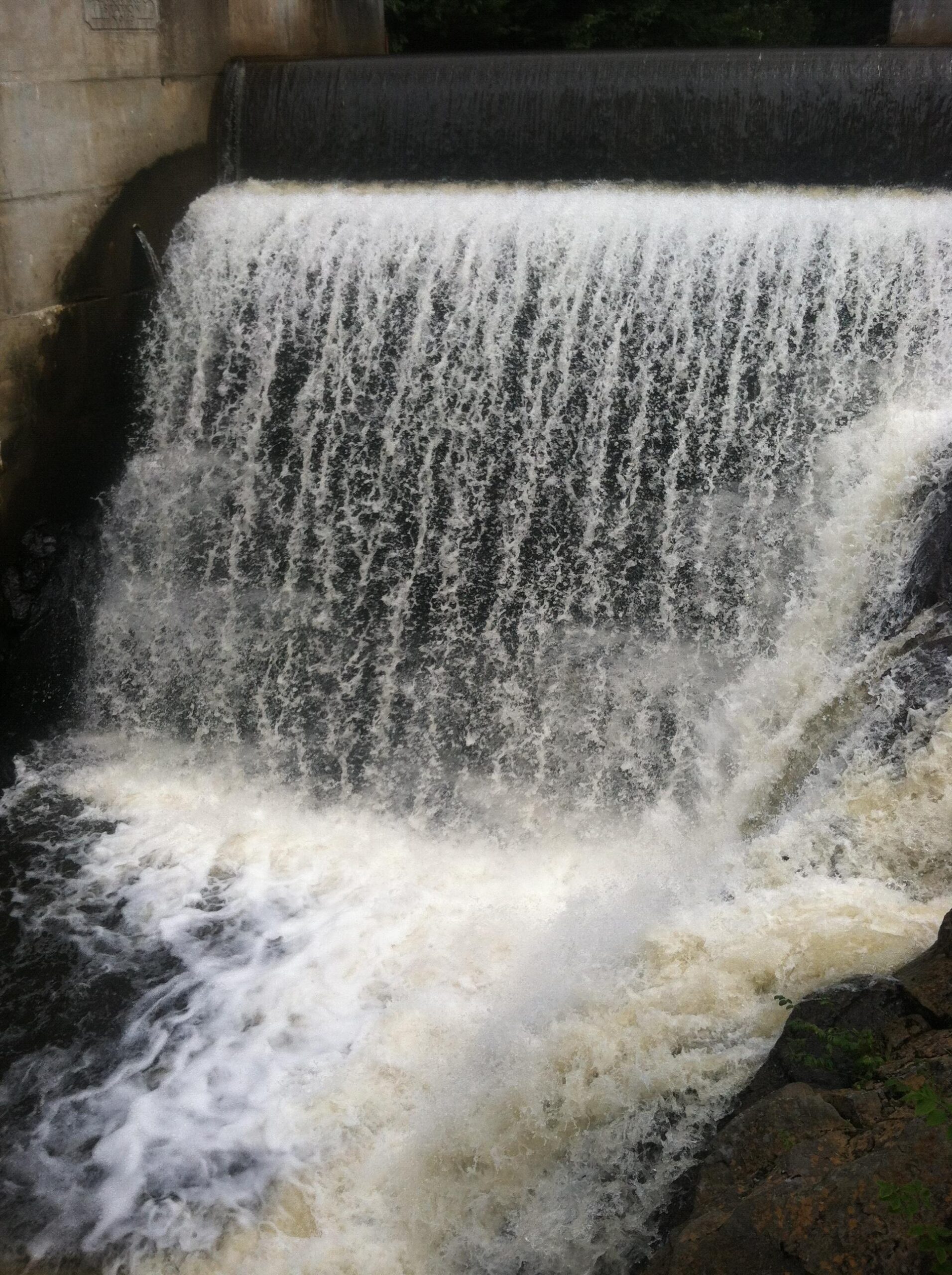 A flowing waterfall cascading over a dam, creating a frothy white spray and foamy water at the base, surrounded by rocky terrain and greenery. Franklin Falls mountain bike trail.