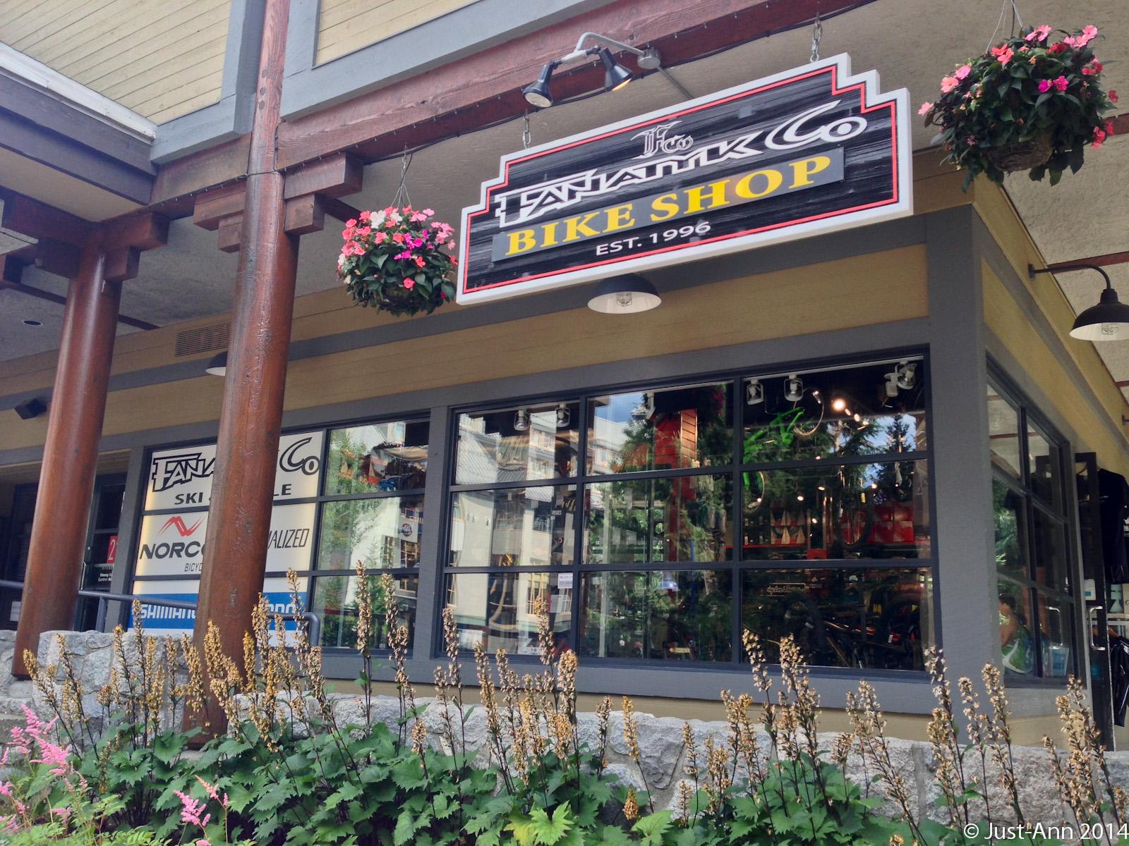 Alt text: Exterior view of a bike shop, featuring a sign that reads "Fantastik Co. Bike Shop" along with hanging flower baskets. The shop's windows display various bicycles and sporting goods, surrounded by flowering plants at the base.