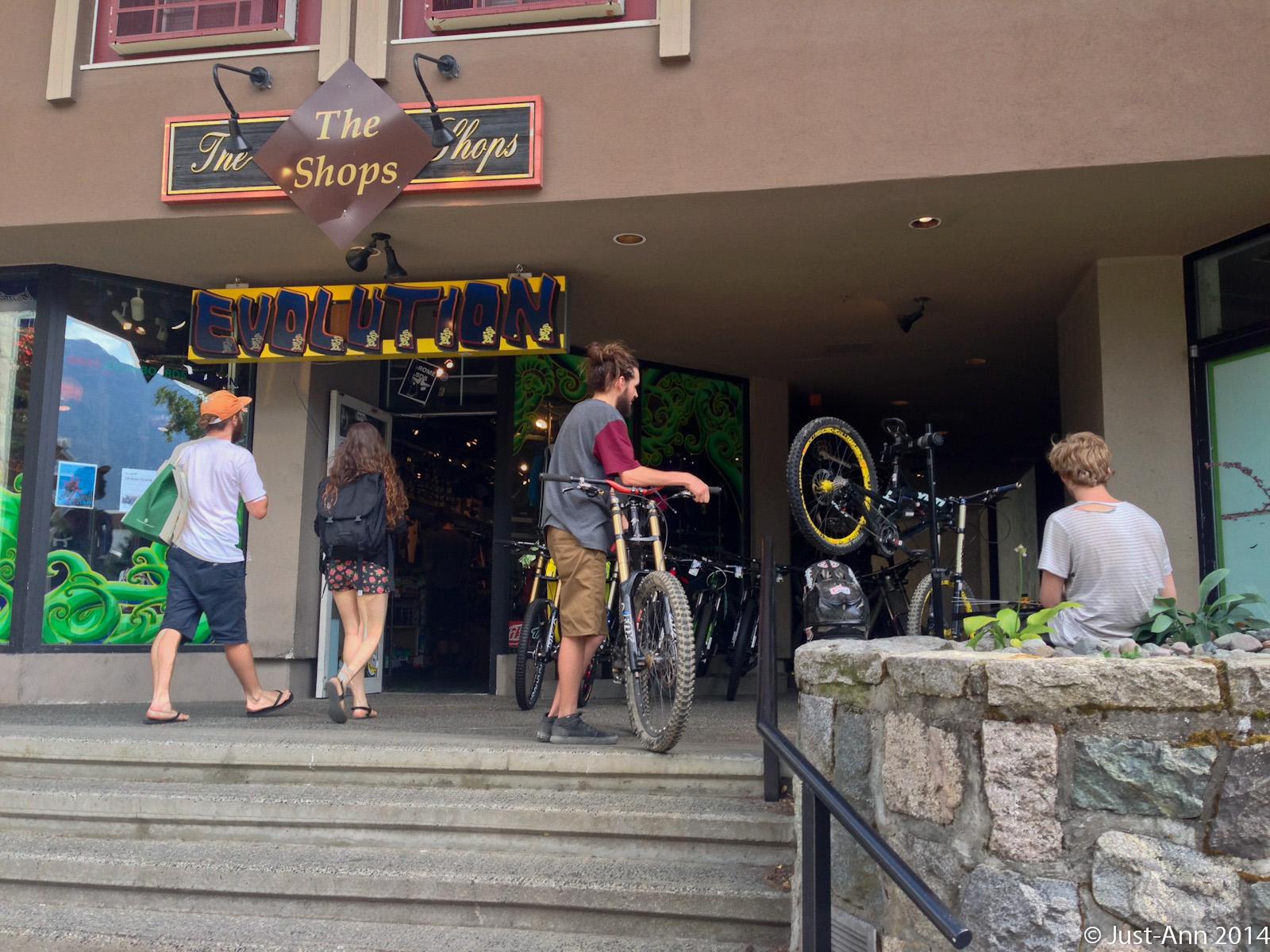 Three individuals are seen outside a shop with the sign "The Shops" and the word "EVOLUTION." Two people are entering the store, one carrying a bag and the other with a backpack. A third person stands on the steps, holding a bicycle, with two additional bicycles visible. The setting appears to be a lively shopping area with greenery in the background.