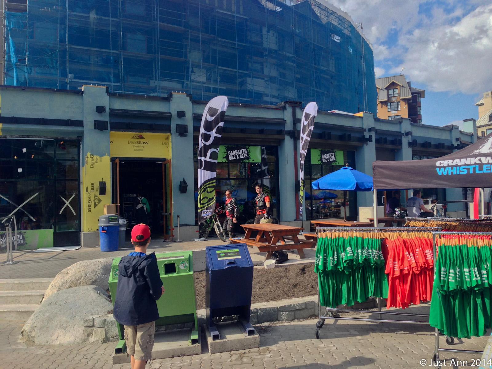 A view of a busy outdoor commercial area featuring a construction site in the background. Two people are seen with bicycles near a storefront, while a child in a red cap stands by recycling bins. In the foreground, a rack of colorful t-shirts in green and red is displayed, and a tent promoting "Crankworx Whistler" is set up nearby. The sky is partly cloudy, suggesting a bright day.
