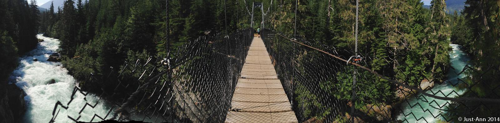 A panoramic view of a suspension bridge spanning a rushing river, surrounded by dense evergreen trees. The bridge features a wooden pathway with a chain-link fence on either side, and a person walking along it. The vibrant blue and white water flows rapidly below, contrasting with the lush greenery of the forest. Cheakamus Lake Trails mountain bike trail.