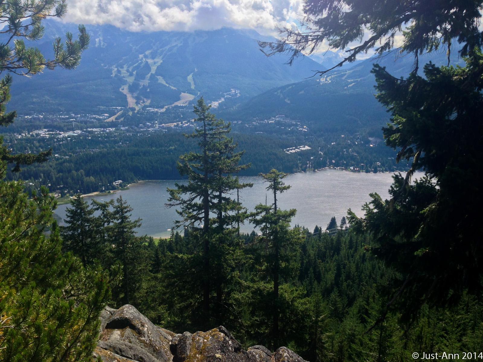 A panoramic view of a mountainous landscape featuring dense evergreen trees in the foreground, a serene lake, and a small town nestled in the valley below, with ski slopes visible on the opposite mountains under a partly cloudy sky. Flank Trail - Function Junction to Rainbow mountain bike trail.