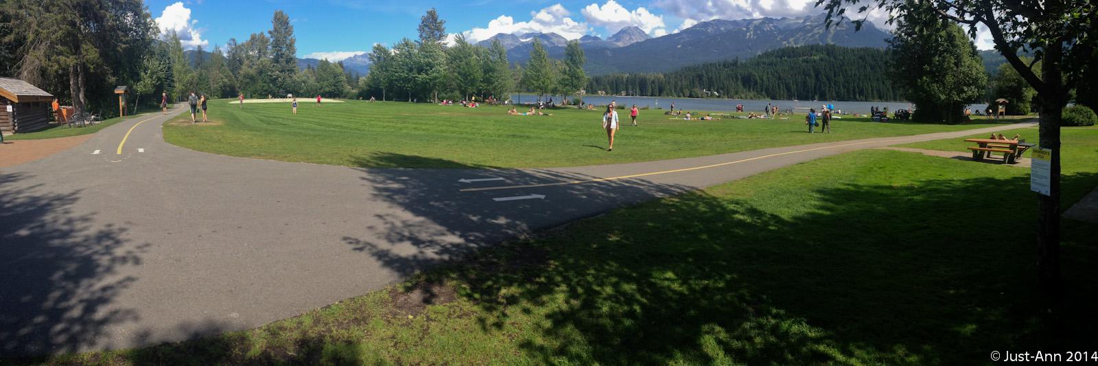 A panoramic view of a sunny park by a lake, featuring green grass, trees, and mountains in the background. People are seen walking or lounging on the grass, while a paved path meanders through the area. There are picnic tables and a log cabin structure on the left side of the image. Bright blue skies with scattered clouds complete the scenic landscape. Whistler Valley Trail mountain bike trail.