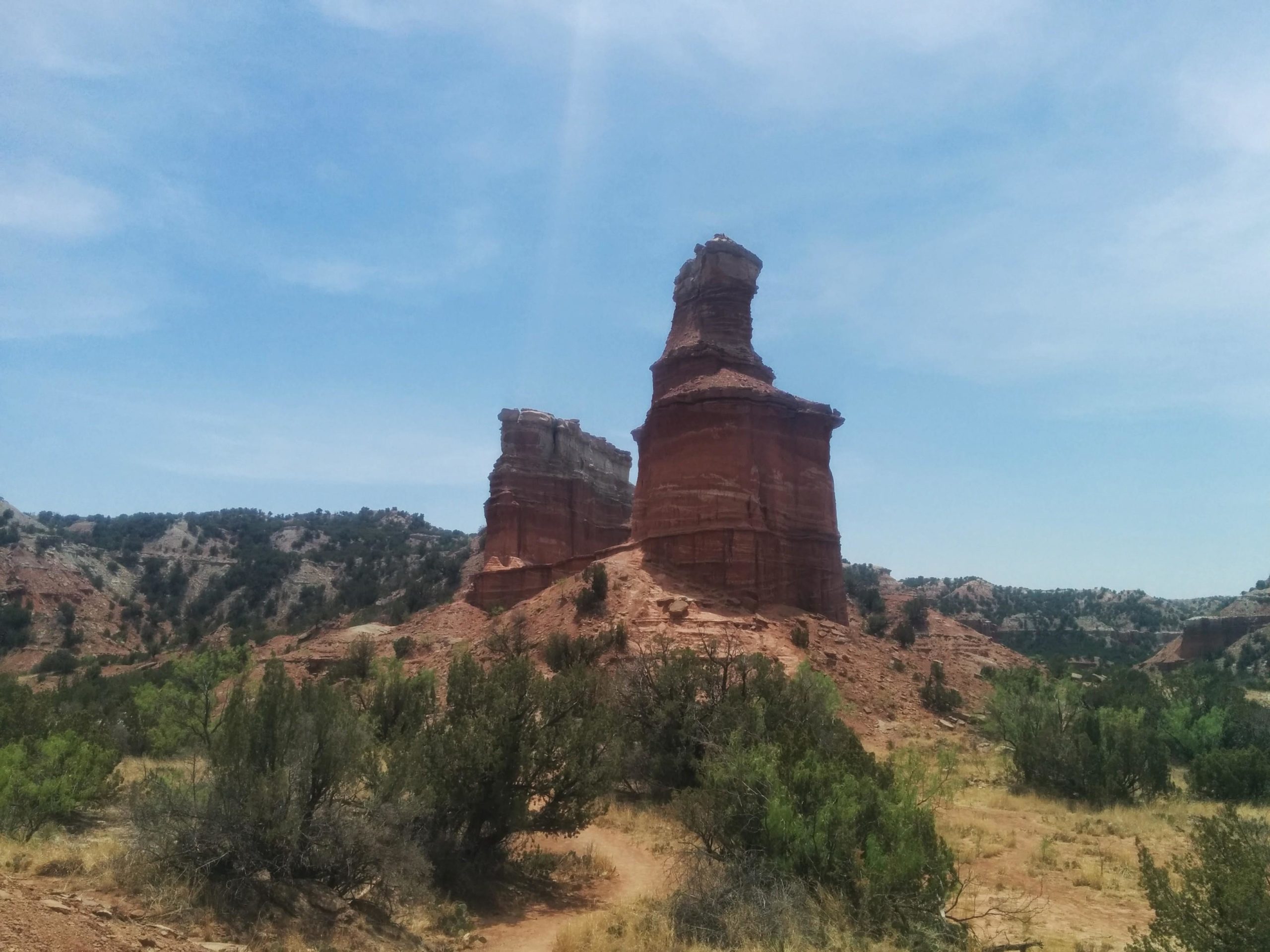 A red rock formation stands prominently against a blue sky, surrounded by rugged terrain and sparse vegetation. The landscape features layered rock structures and distant hills, capturing the natural beauty of a desert or canyon environment. Palo Duro Canyon mountain bike trail.
