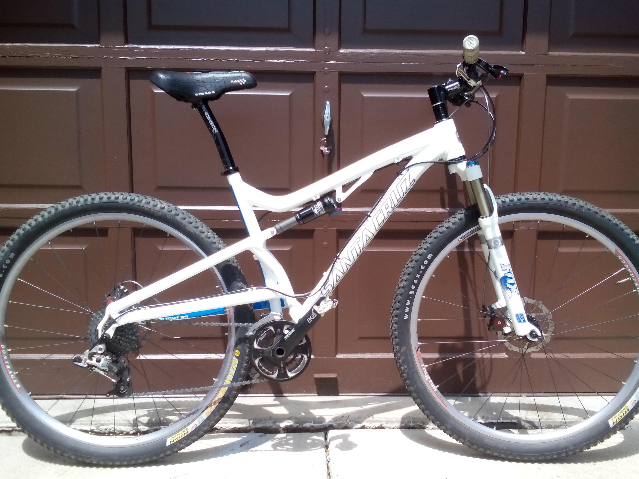 Santa Cruz SuperLight 29: A white mountain bike with black and blue accents is leaning against a brown garage door. The bike features a dual-suspension frame, knobby tires, and a prominent front fork. The seat is raised, and the gear mechanism is clearly visible, showcasing its design for off-road terrain.