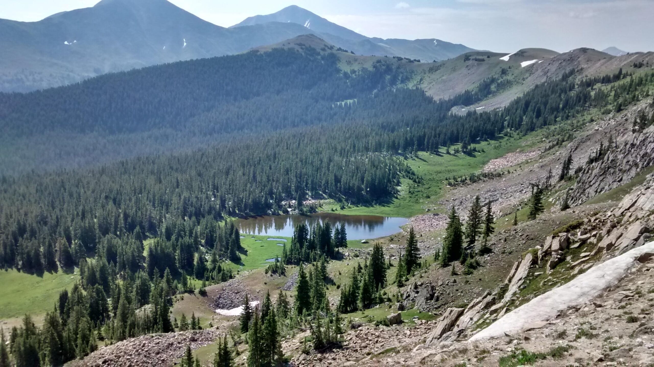 A panoramic view of a lush green valley surrounded by mountains, featuring a small lake nestled among dense forests of evergreen trees. The landscape is characterized by rocky terrain on the right side, with sunlight illuminating the scene under a clear sky. Monarch Crest Trail mountain bike trail.