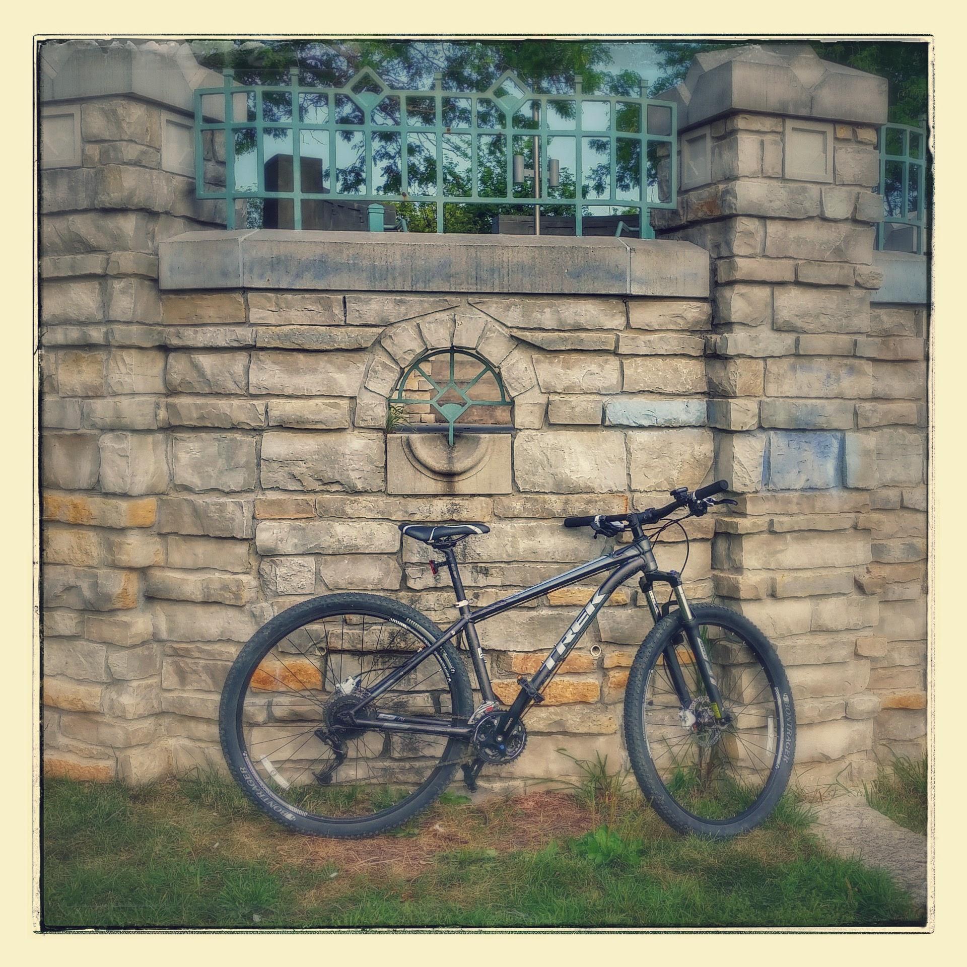 Trek X caliber 6: A black mountain bike leaned against an elaborately designed stone wall with a circular emblem, surrounded by grass. The wall features a green iron gate and decorative stonework.