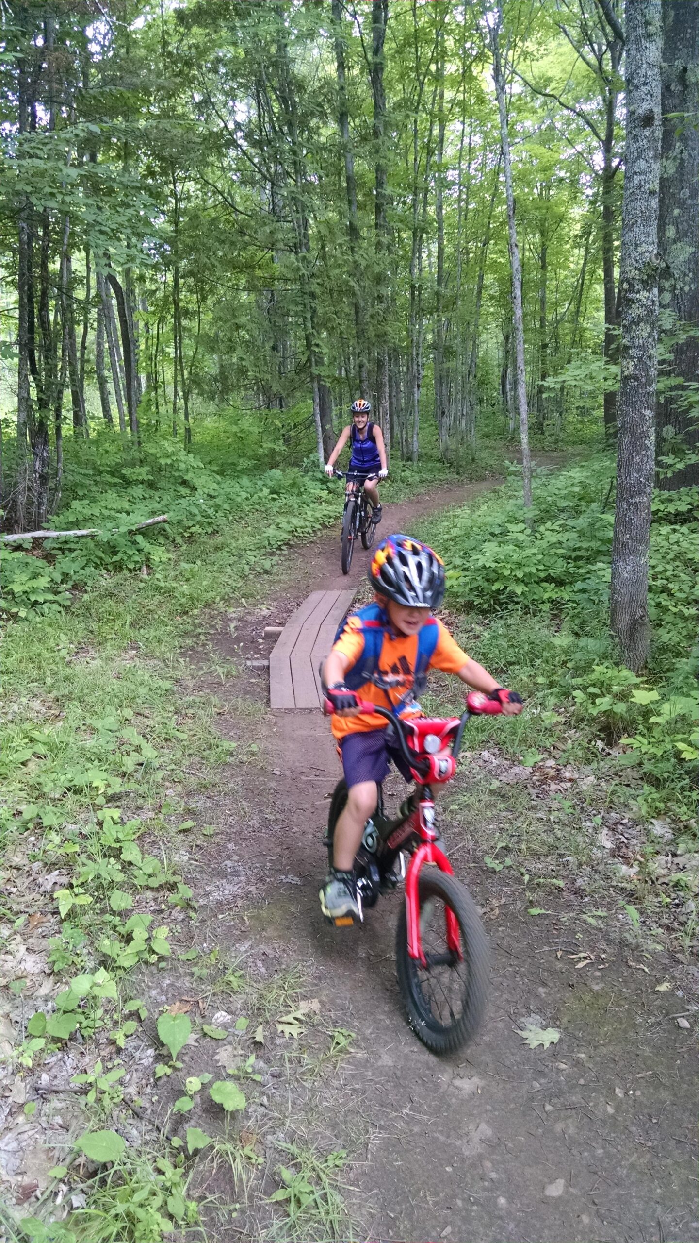 A child riding a small red bicycle along a dirt trail in a wooded area, while an adult on a mountain bike follows behind. The scene is surrounded by lush green trees and foliage, creating a vibrant and natural setting. The child is wearing a helmet and bright orange shirt, while the adult is dressed in a blue tank top with a black backpack. Michigan Tech Trails mountain bike trail.