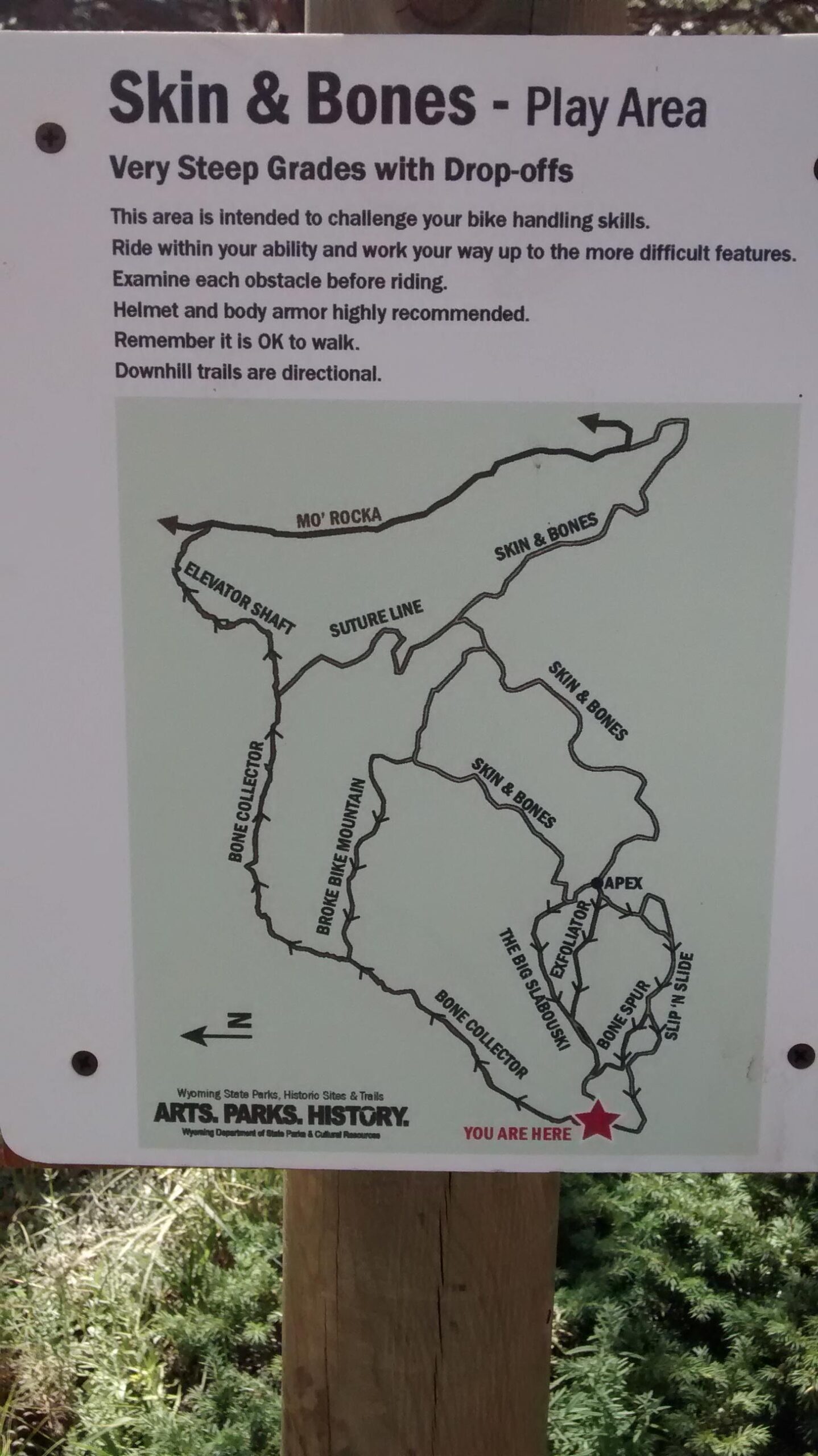 Sign for the "Skin & Bones" play area, featuring bike trails with steep grades and drop-offs. The sign includes safety recommendations, a reminder to examine obstacles, and indicates that helmets and body armor are recommended. It also shows a map of the trails, highlighting various routes and indicating the viewer's location with a star. Curt Gowdy State Park mountain bike trail.