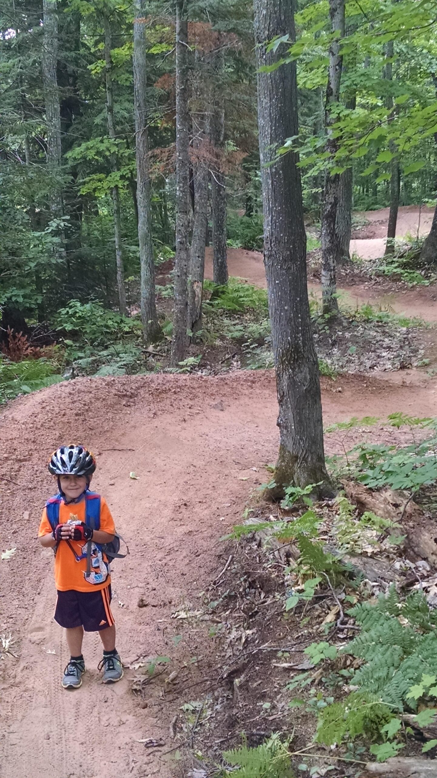 A young boy wearing a helmet and an orange shirt stands on a dirt trail in a wooded area, holding a camera. The trail winds through trees and greenery, indicating a biking or hiking path. Michigan Tech Trails mountain bike trail.