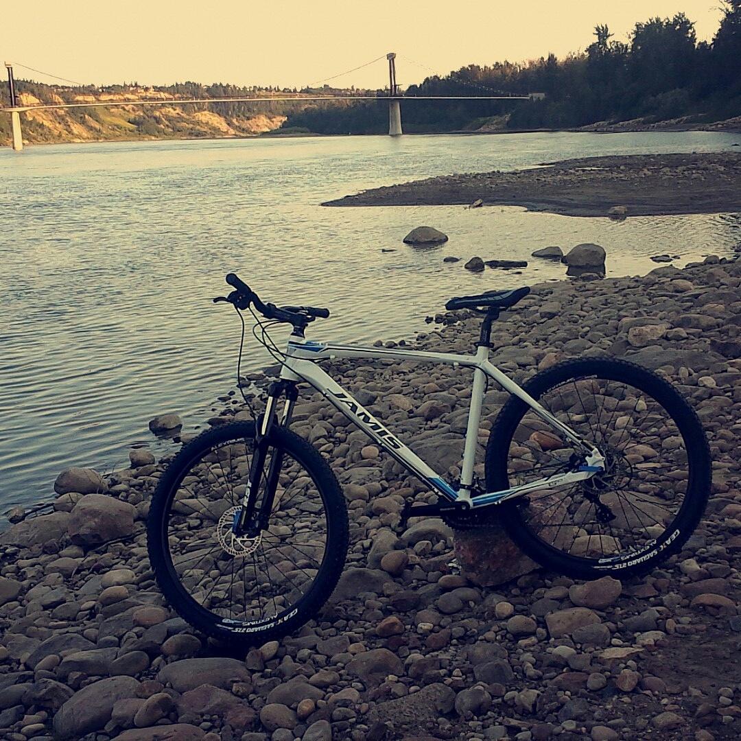 Jamis Nemesis: A mountain bike is parked on a rocky shoreline beside a calm river, with a bridge visible in the background and trees lining the opposite bank. The scene is illuminated by soft, warm light, suggesting early evening.