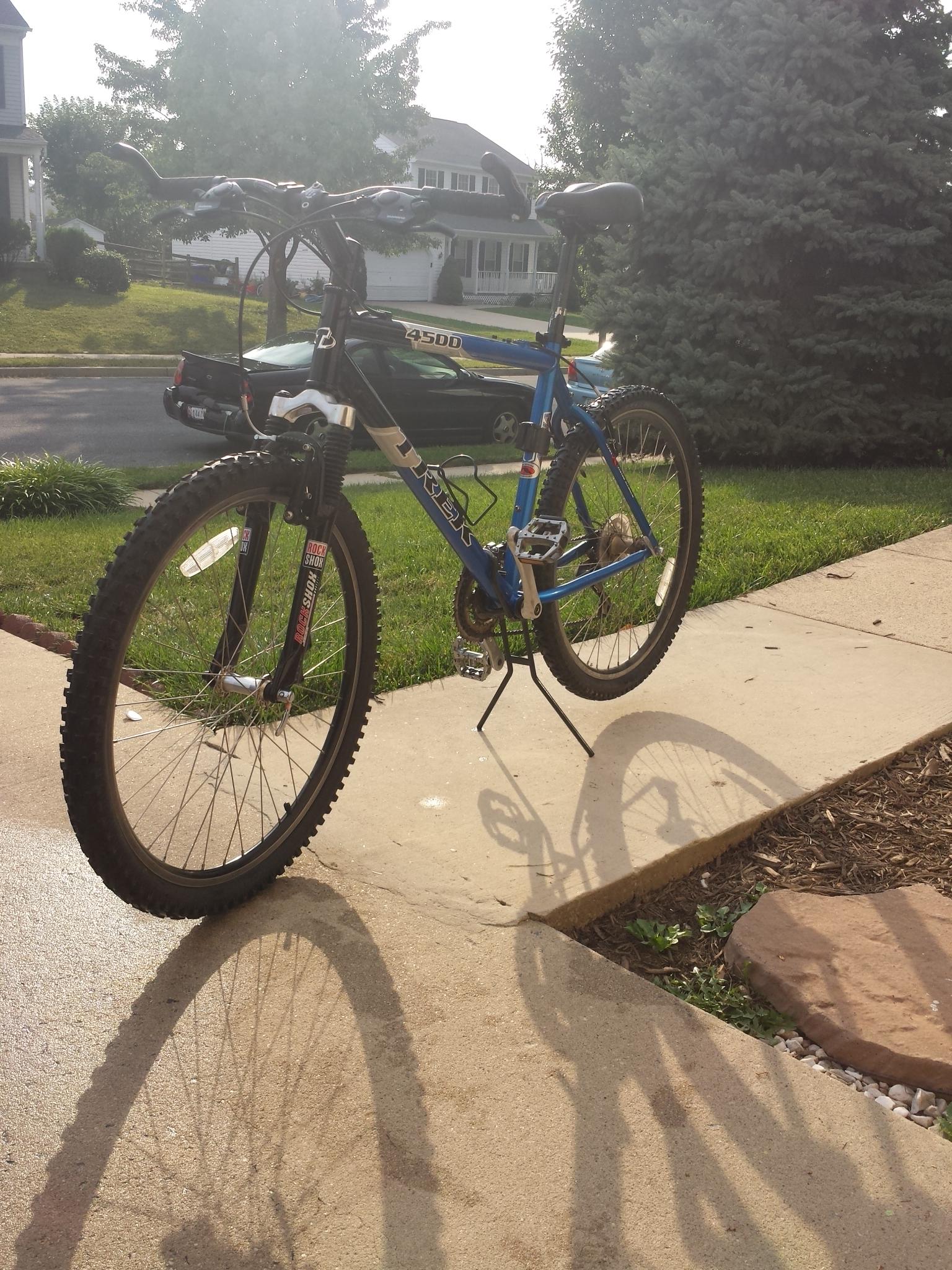 Trek 4500: A blue mountain bike on a concrete sidewalk, casting a shadow. The bike is positioned beside a grassy area and a decorative stone. The background features houses, trees, and a parked car, with morning sunlight illuminating the scene.