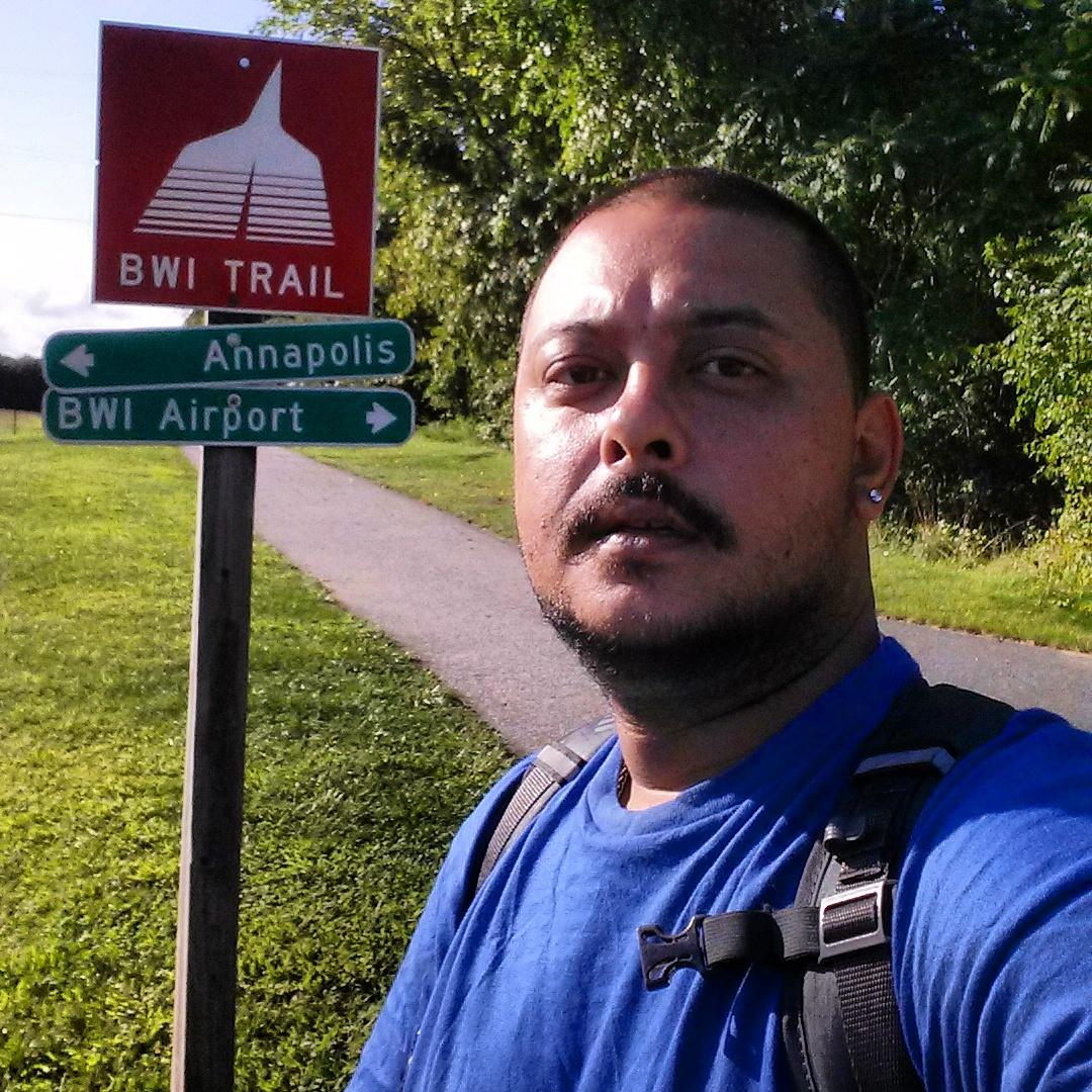 Trek 4500: Person standing in front of a sign indicating directions to Annapolis and BWI Airport, with a background of greenery and a walking path. The individual is wearing a blue shirt and has a backpack.