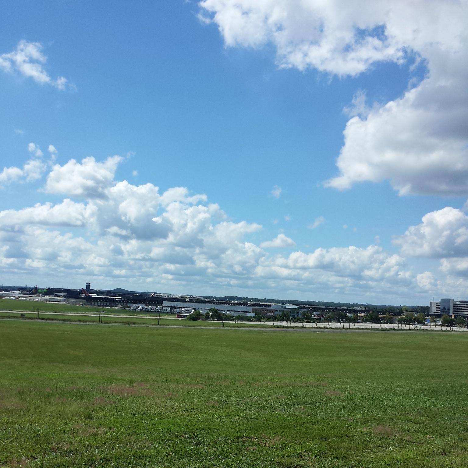 Trek 4500: A clear blue sky dotted with fluffy white clouds above a grassy field, with an airport visible in the background, featuring multiple airplanes and terminal buildings.