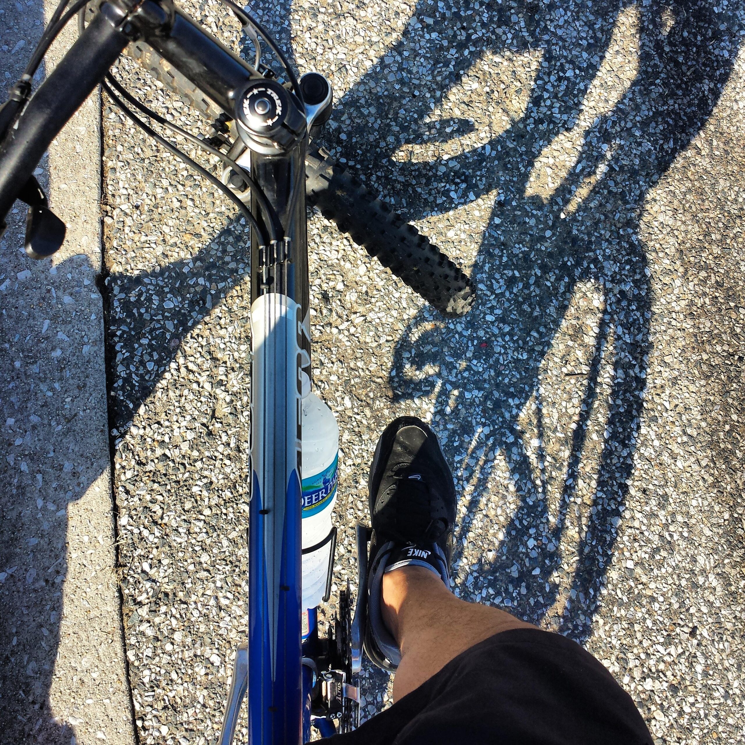 Trek 4500: A top-down view of a cyclist's foot resting on a bike pedal, with a water bottle attached to the bike frame. The background shows a textured asphalt surface, and the shadows of the bike and rider are visible on the ground.