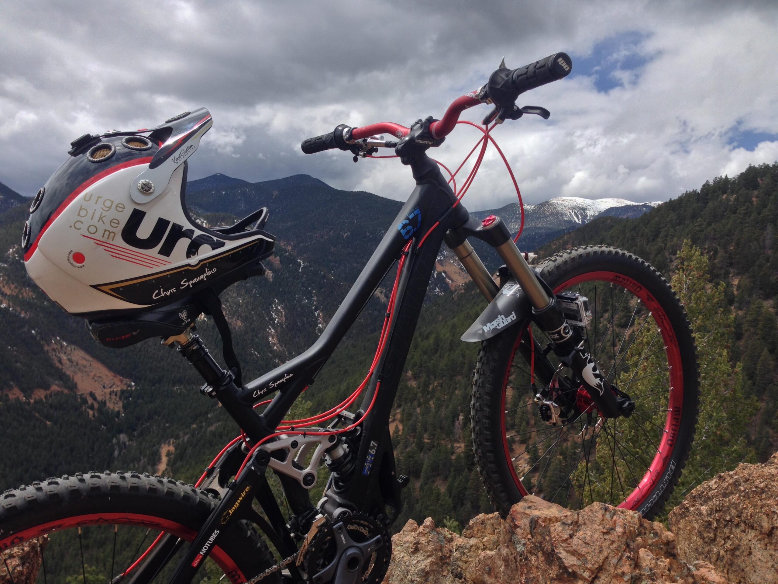 Giant Reign X1: Mountain bike leaning against a rocky outcrop with a helmet on the seat, surrounded by a scenic mountainous landscape under cloudy skies.