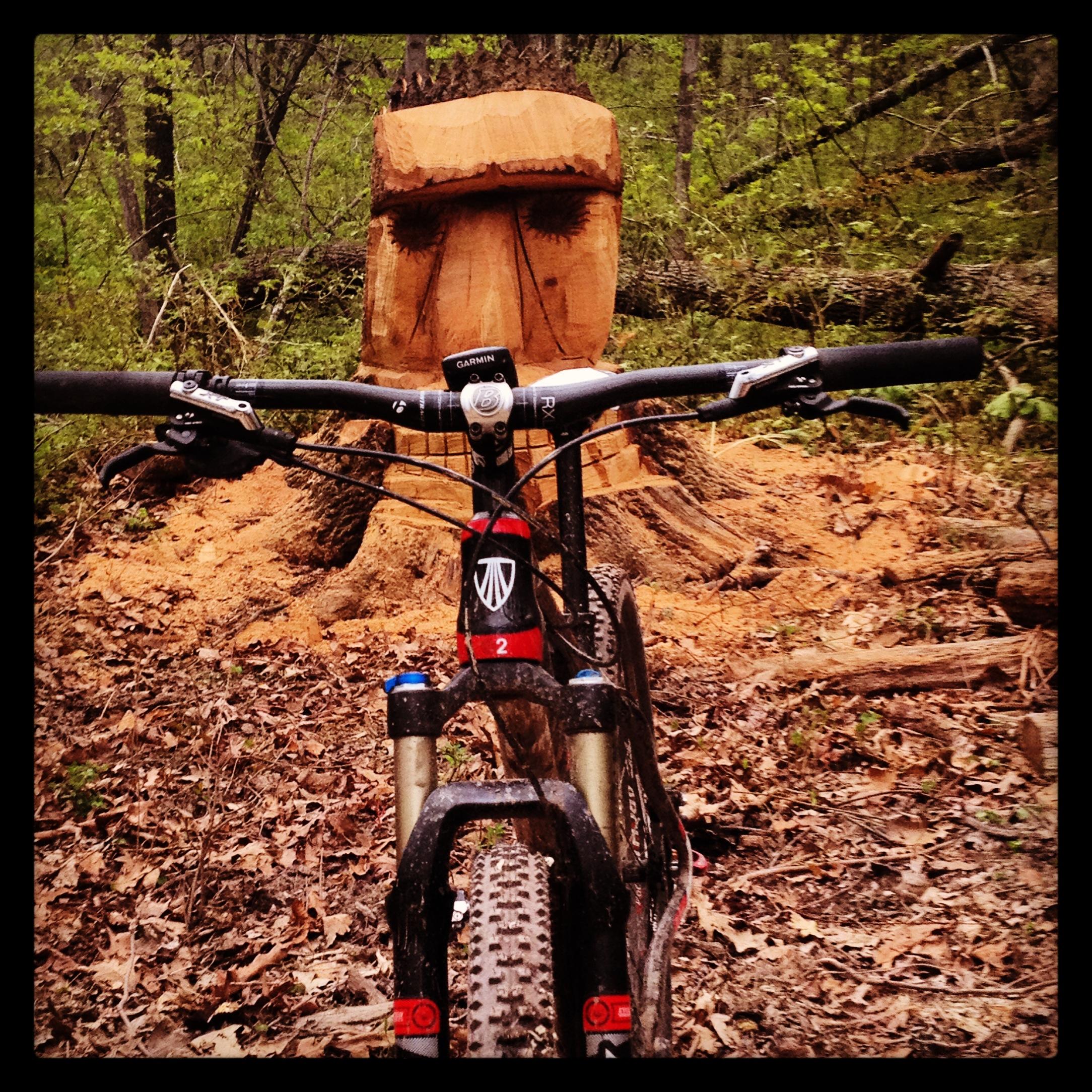Alt tag: "Mountain bike positioned in front of a carved wooden face sculpture, surrounded by forest foliage and fallen leaves." Nockamixon State Park mountain bike trail.