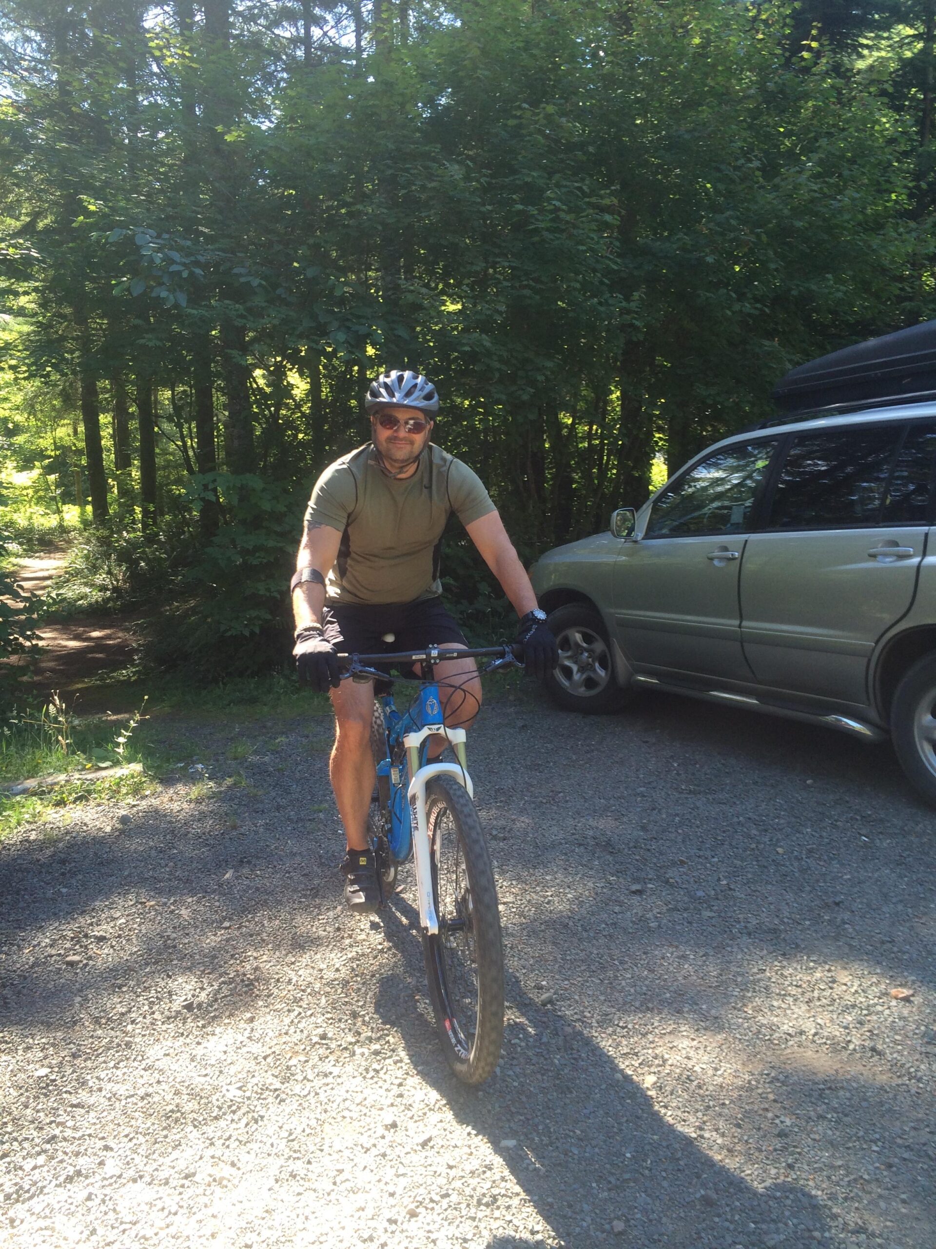 Niner RIP 9: A person in a helmet and sunglasses rides a mountain bike on a gravel path, surrounded by dense greenery. A parked vehicle is visible in the background, partially obscured by trees.