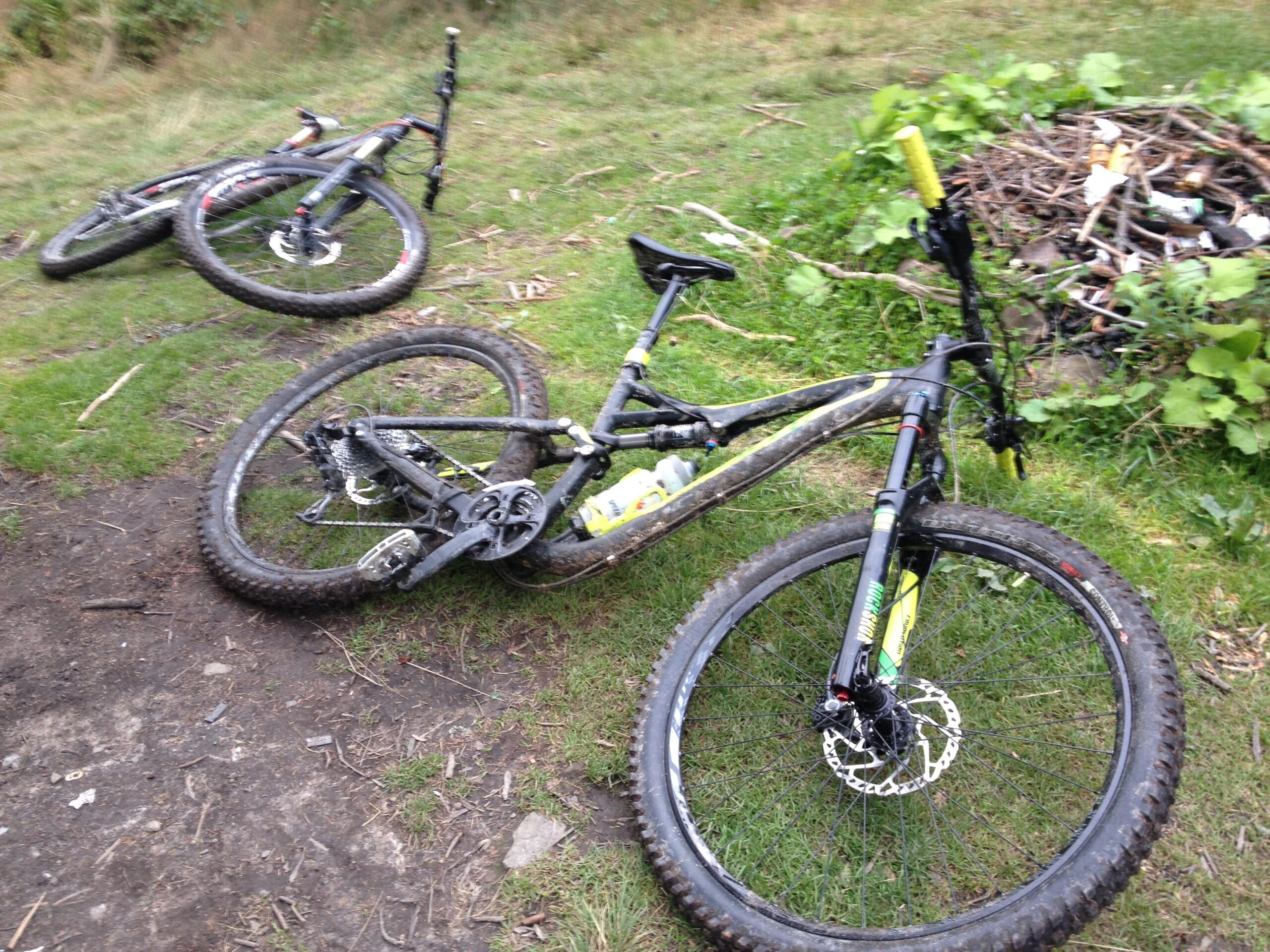 Specialized Stumpjumper FSR: Two mountain bikes lying on the grass, partially covered in mud, near a small pile of sticks and debris. The bikes are positioned at an angle, with one in the foreground and the other slightly behind it. Surrounding vegetation includes patches of grass and some leafy plants.