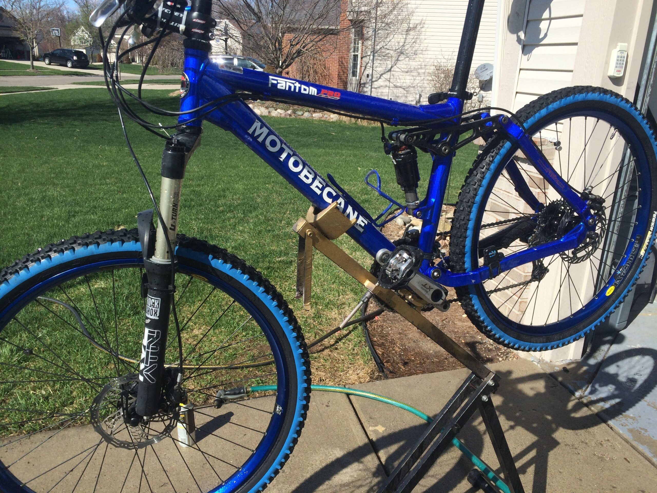 Motobecane Fantom PRO Titanium: A blue mountain bike with a prominent "Motobecane" label is positioned on a bike stand for maintenance. The bike features thick, knobby tires with blue rims, a front suspension fork, and visible gears and brakes. The background includes a grassy area and a residential setting, indicating an outdoor environment.