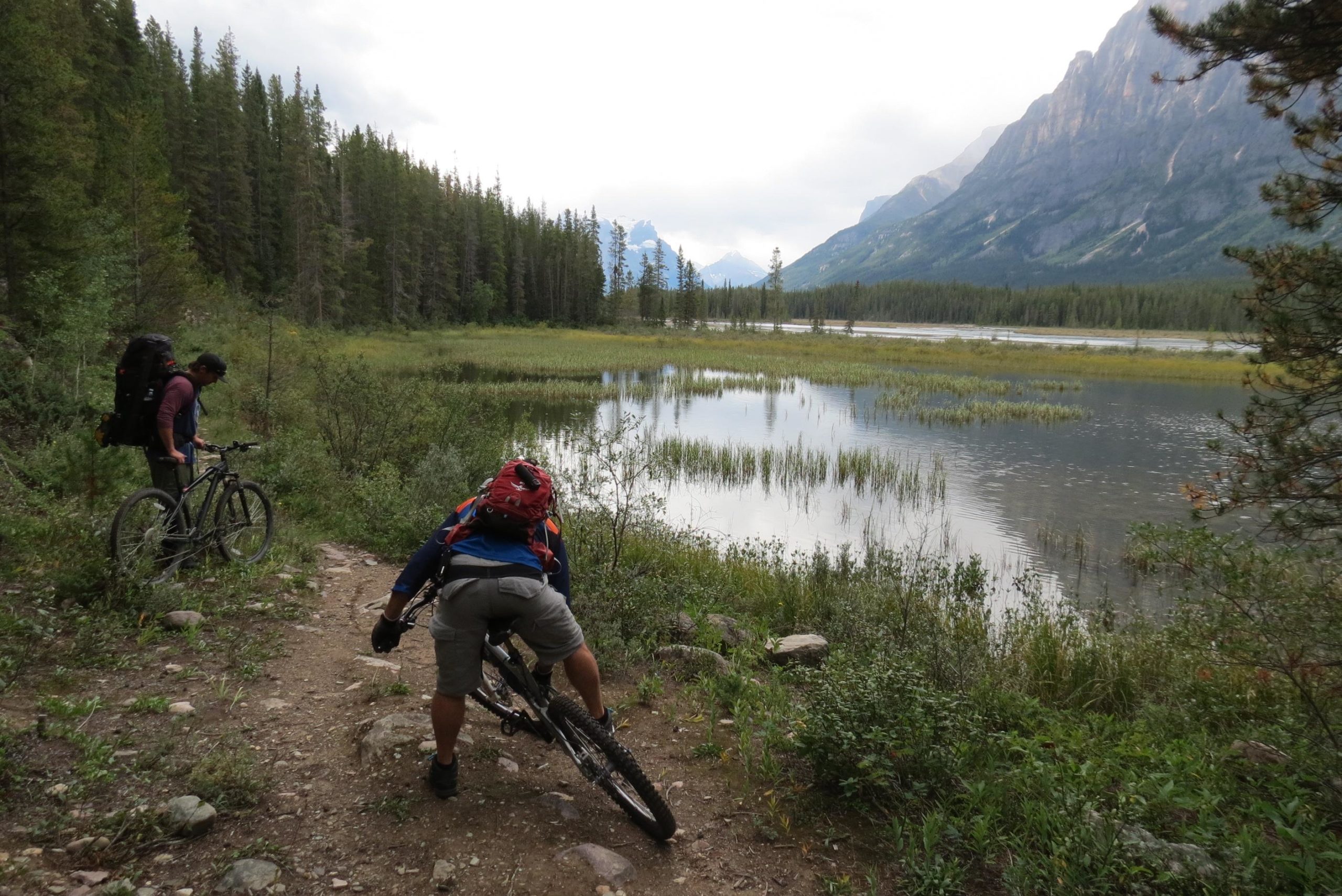 Two mountain bikers navigating a trail near a body of water, surrounded by tall trees and mountains in the background. One biker is adjusting his bike while the other appears to be preparing to ride. The scene captures a serene outdoor environment, with a mix of greenery and a reflective water surface. Big Bend (Trail #126) mountain bike trail.