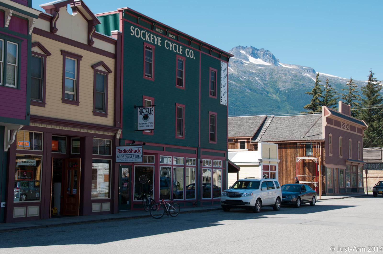 A vibrant street scene featuring colorful historic buildings, including a green shop labeled "Sockeye Cycle Co." and a "RadioShack" storefront. The background showcases majestic mountains with snow-capped peaks and clear blue skies. Parked vehicles and a bicycle are visible along the street.