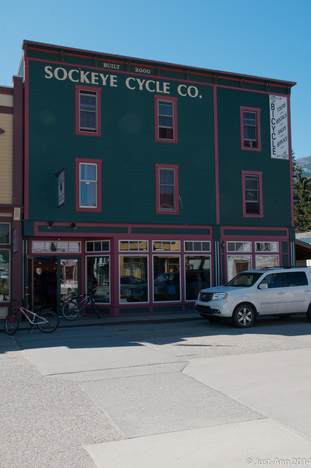 A green two-story building with red trim featuring the sign "Sockeye Cycle Co." on the front. The shop offers bicycle rentals, tours, sales, and service. Bicycles are parked outside, and a white SUV is visible in front of the store. Clear blue skies are overhead.