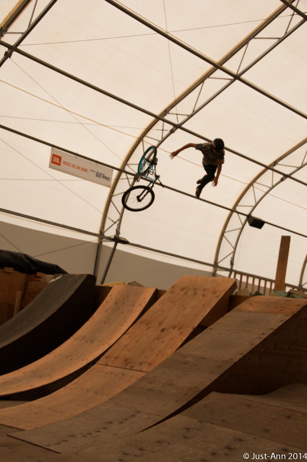 A BMX rider performing a trick in mid-air above a series of wooden ramps inside a large indoor skate park. The rider is in a dynamic pose, with one hand on the bike, while the bike is positioned close to them. The tent-like structure overhead is visible, providing a spacious environment for action sports. Whistler Air Zone mountain bike trail.