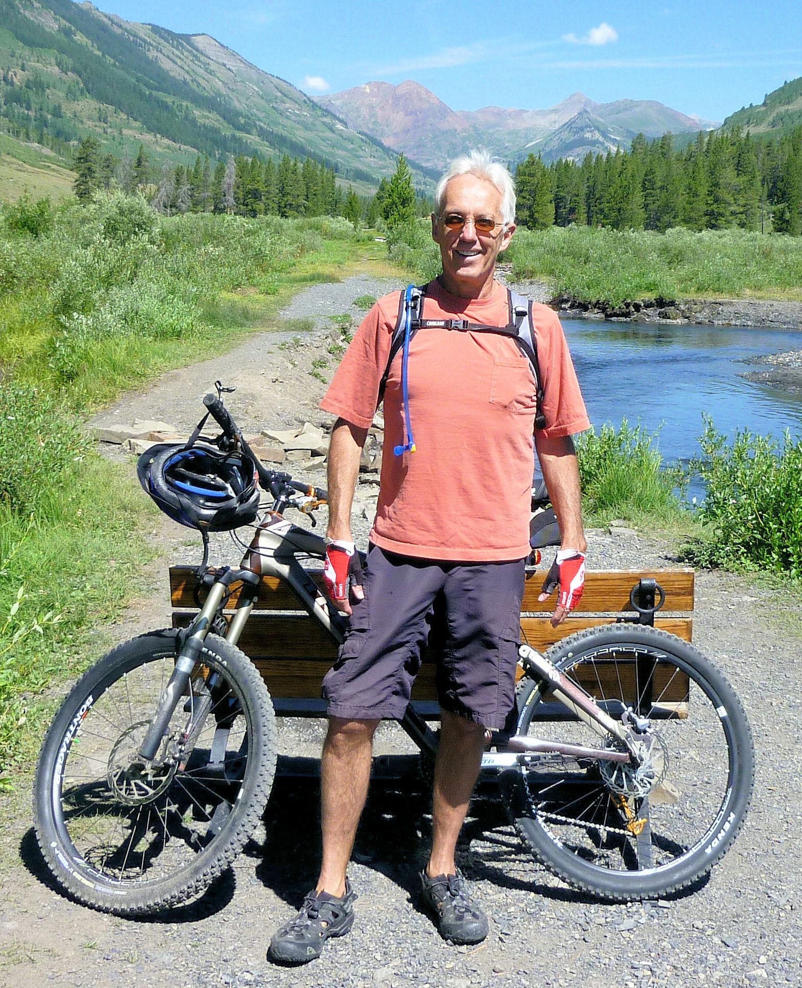 Trek Remedy 9.9: An older man stands next to a mountain bike on a gravel path, surrounded by lush greenery and mountains in the background. He is wearing a bright orange shirt, shorts, and gloves, and has a hydration pack on his back. A helmet rests on the bike's handlebars. The scene is sunny with a clear blue sky.