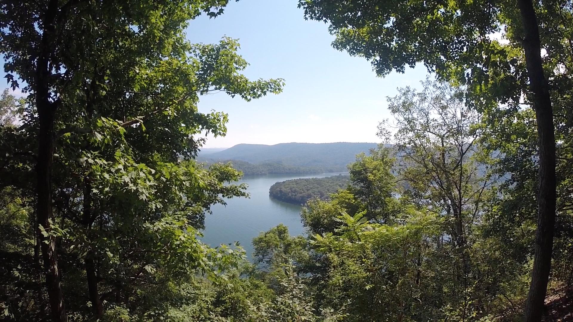 A scenic view of a calm lake surrounded by lush green trees and rolling hills under a clear blue sky. The image captures a natural landscape with vibrant foliage framing the water and distant mountains. Allegrippis Trails mountain bike trail.