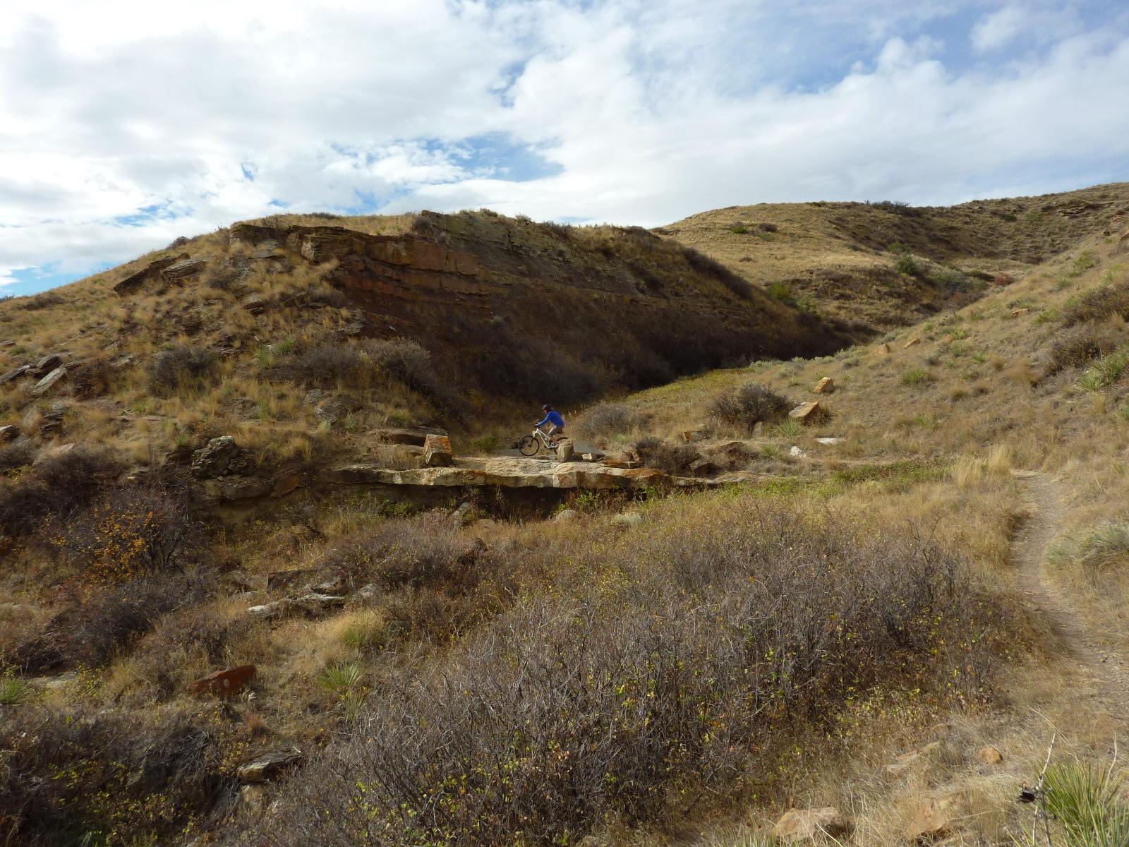 A cyclist navigating a rocky terrain surrounded by dry grass and shrubs, with rolling hills in the background under a partly cloudy sky. North Shore mountain bike trail.
