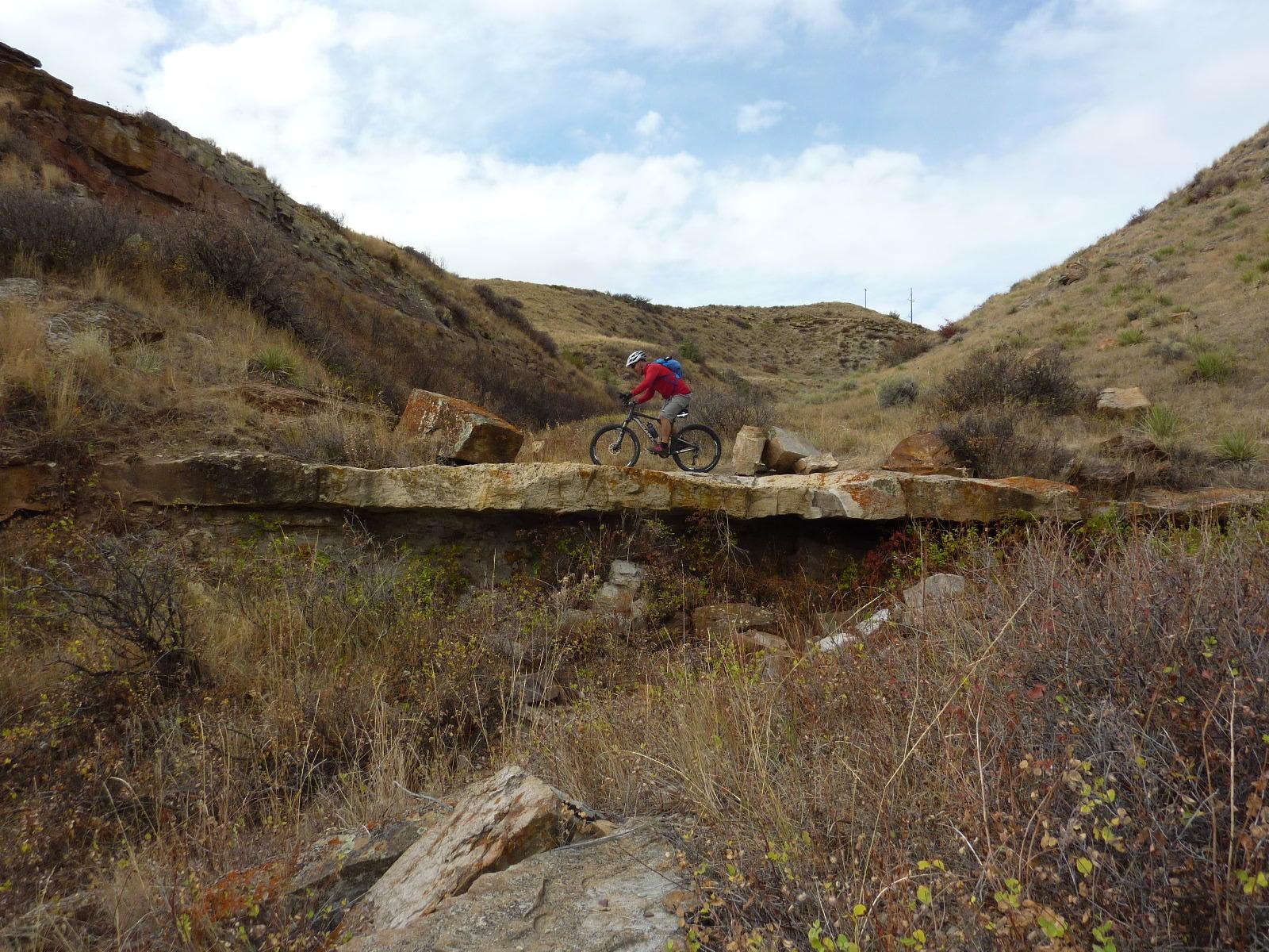 A mountain biker gracefully navigates a rocky terrain while riding over a stone bridge in a hilly landscape, surrounded by dry grass and sparse vegetation under a partly cloudy sky. North Shore mountain bike trail.