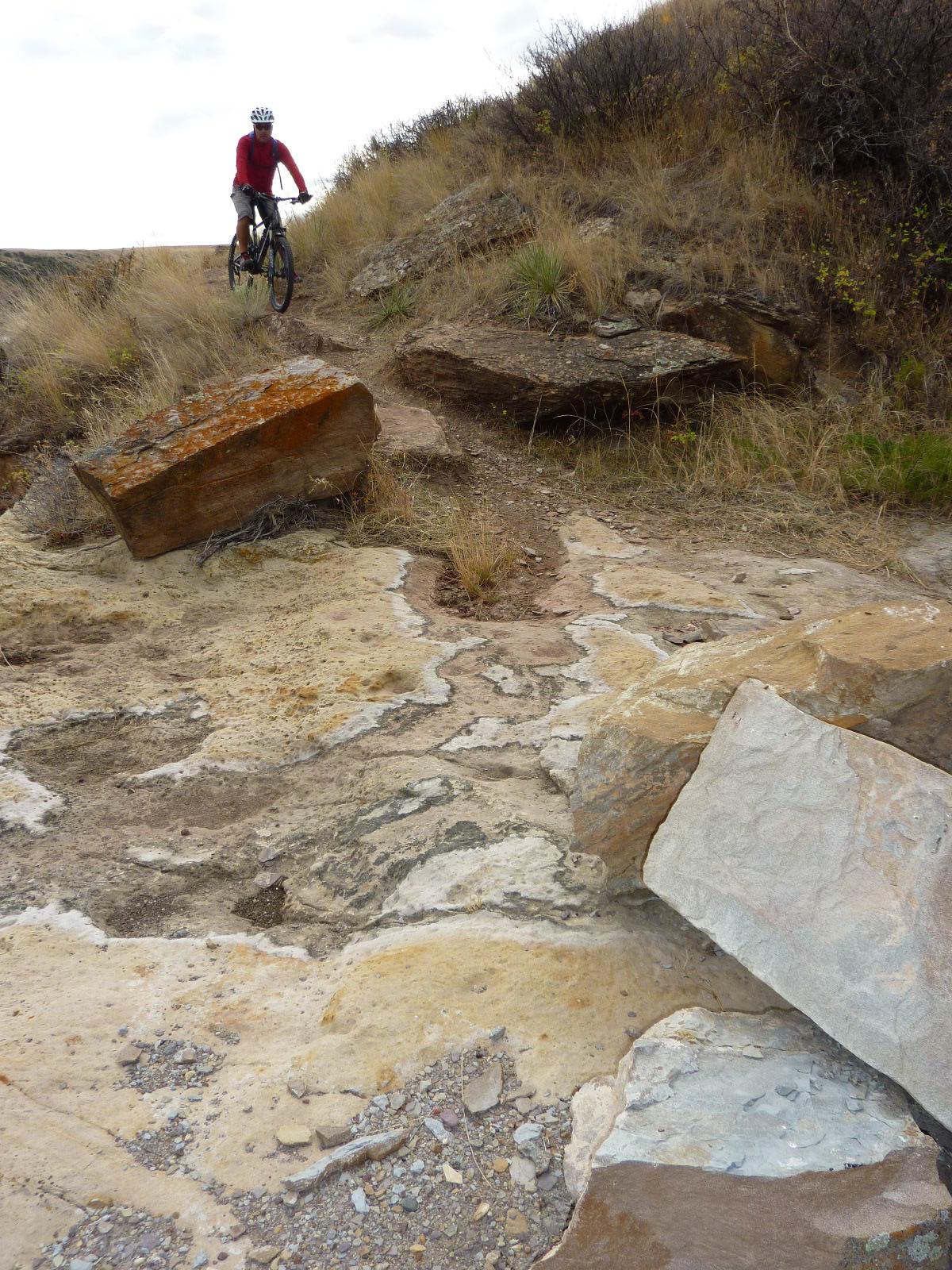 A person riding a mountain bike on a rocky trail with large boulders and sparse vegetation in a natural outdoor setting. The terrain is rugged, with patches of dirt and stone, and the sky is overcast. North Shore mountain bike trail.