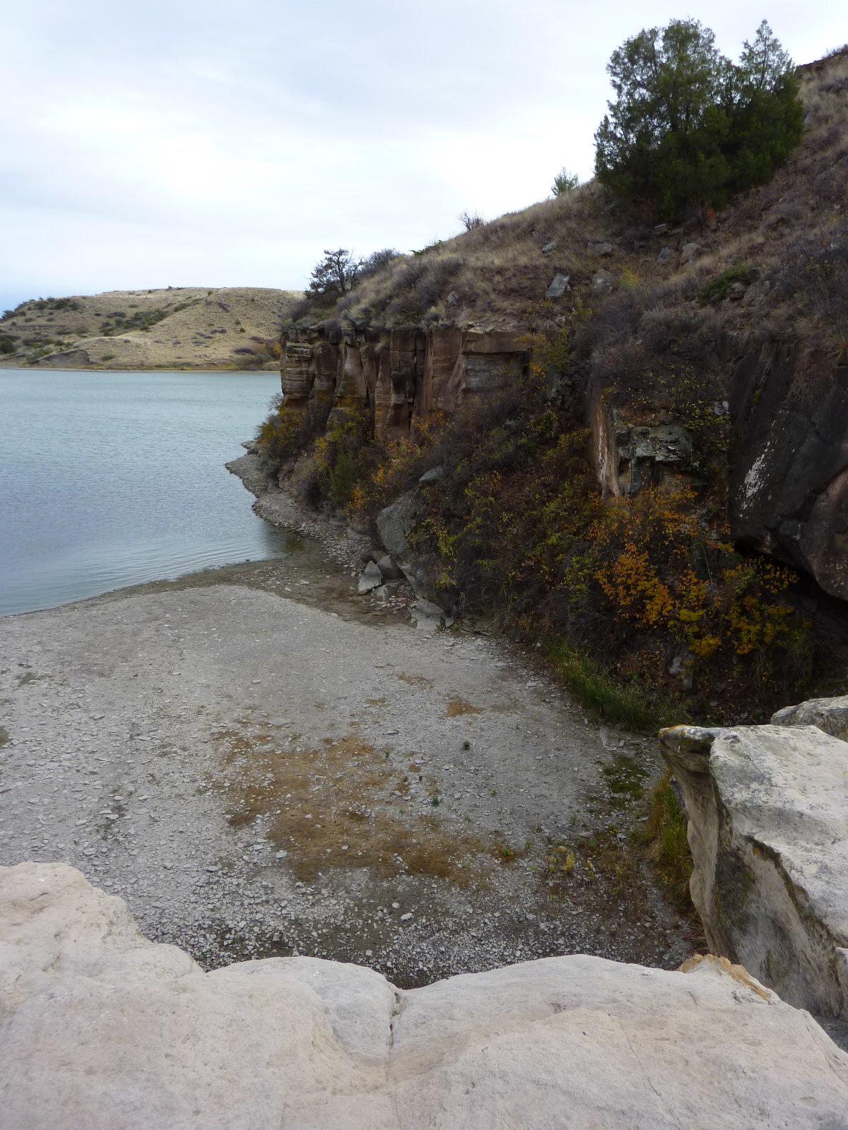 A scenic view of a lake bordered by rocky cliffs and sloped terrain. The shoreline features a mix of gravel and sparse vegetation, with some autumn-colored foliage visible. In the background, rolling hills rise against a cloudy sky, creating a tranquil natural landscape. North Shore mountain bike trail.