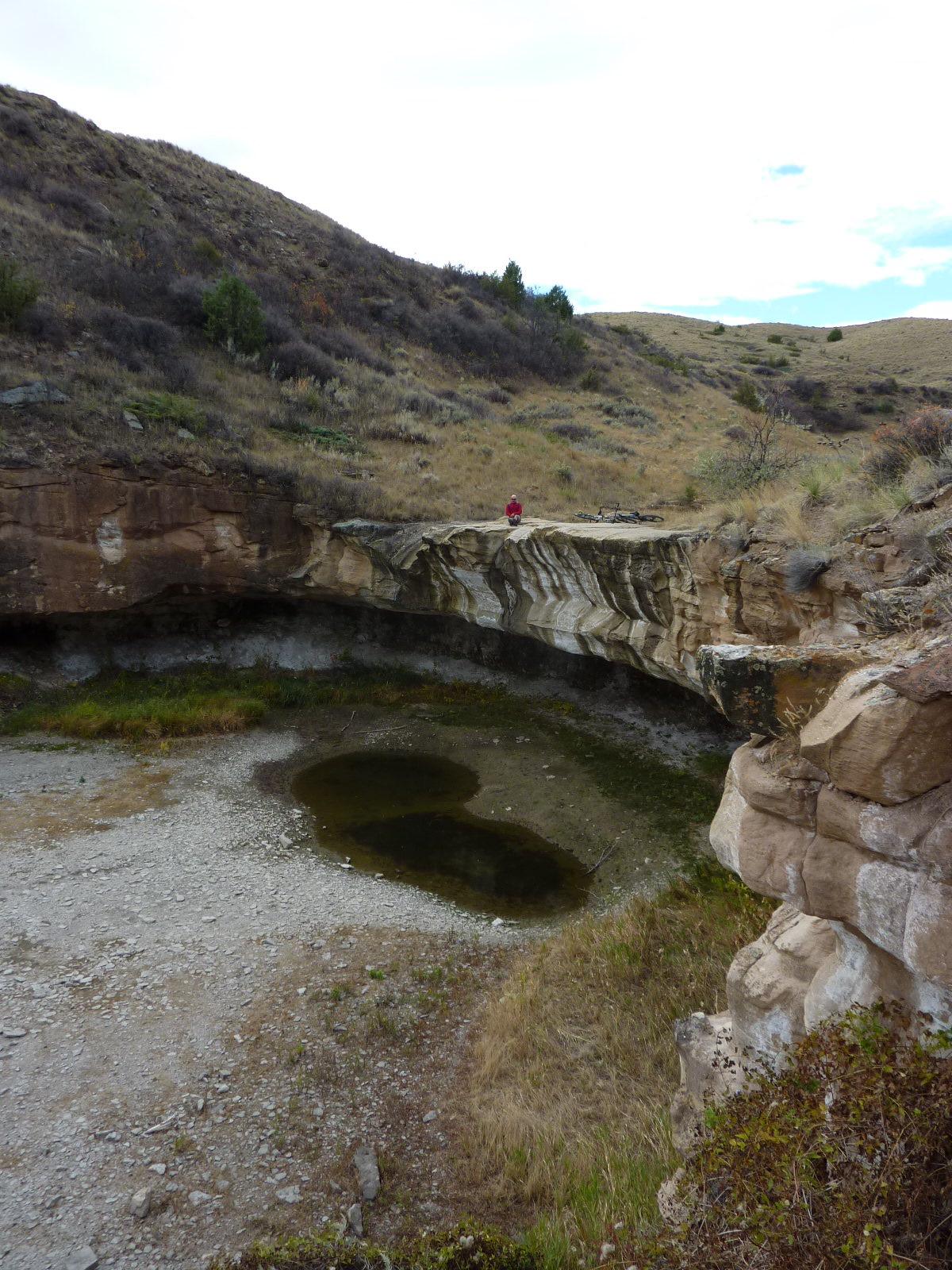 A person stands on the edge of a rocky ledge overlooking a small, shallow water pool surrounded by beige and gray rock formations. The landscape features rolling hills with sparse vegetation, including patches of greenery and shrubs. The sky is partly cloudy, creating a serene natural setting. North Shore mountain bike trail.