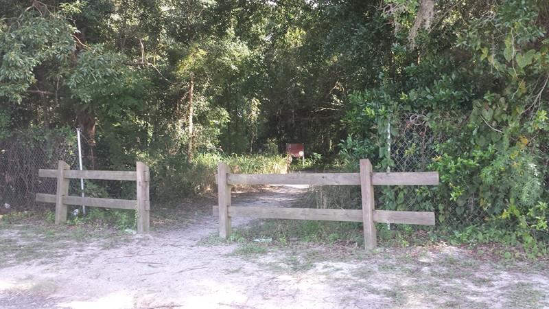 A wooden fence encloses an overgrown pathway leading to a dense tree line. The path is sandy and unpaved, bordered by foliage on either side, with a glimpse of a sign visible in the background among the trees. The area is shaded by lush greenery, suggesting a natural setting. Mount Dora Trail mountain bike trail.