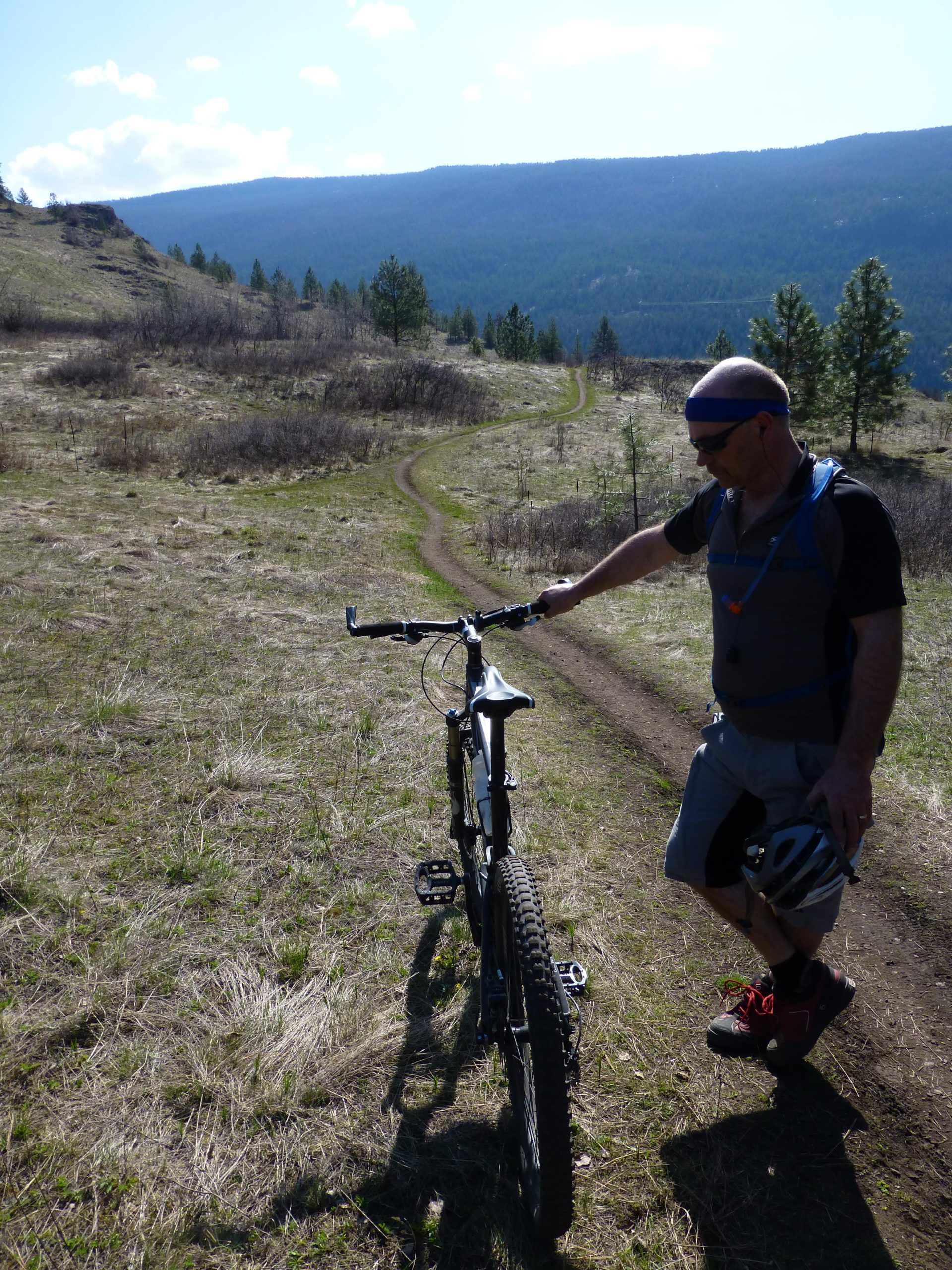 A mountain biker stands beside his bike on a grassy trail with a winding path in the background. The landscape features rolling hills and a mountain range under a clear blue sky. The cyclist is wearing a helmet and sports attire, looking down at the bike while catching his breath. Kalamalka Lake Provincial Park mountain bike trail.