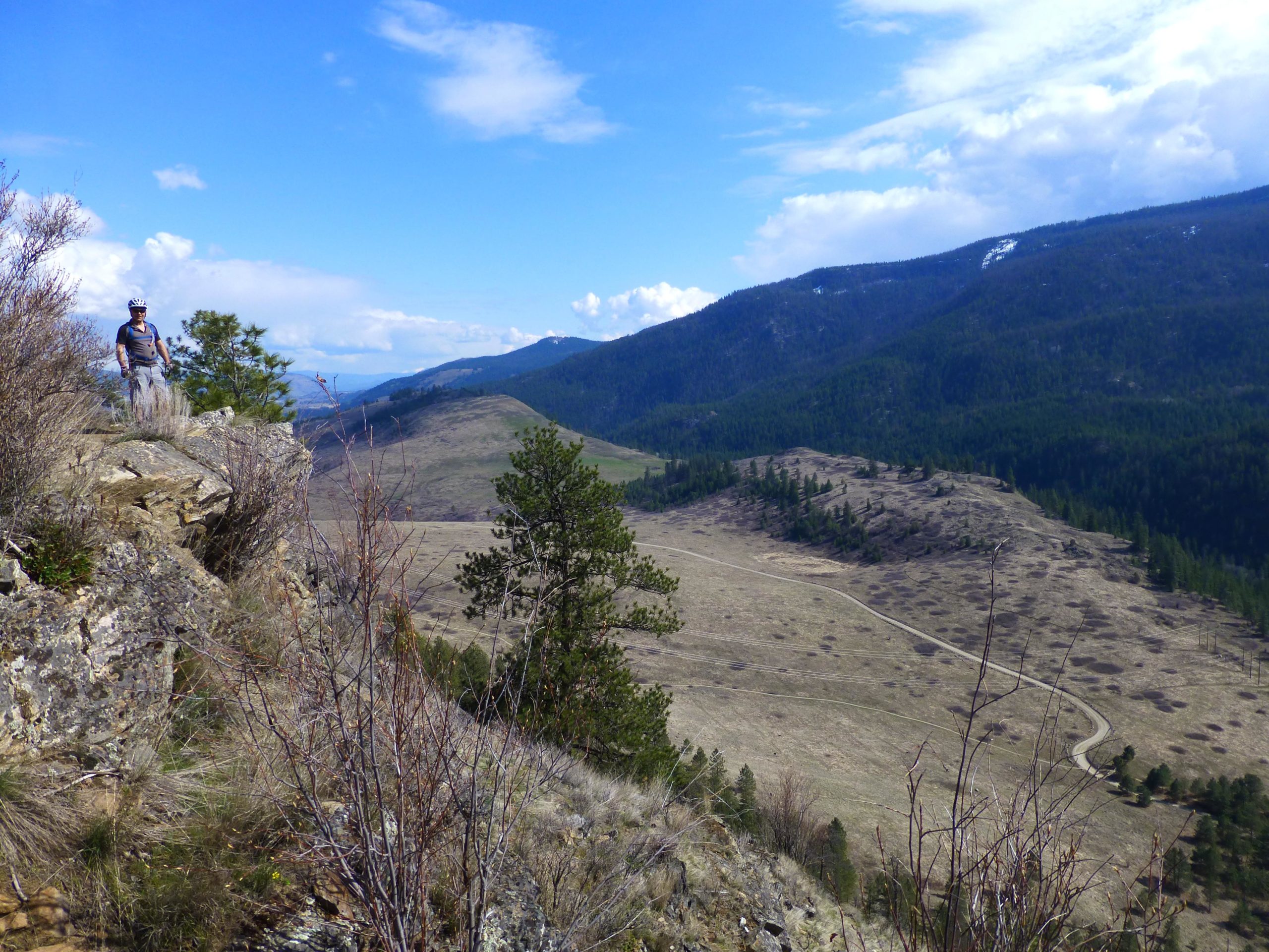 A person standing on the edge of a rocky outcrop, overlooking a wide valley with rolling hills and scattered trees. The sky is blue with some clouds, and snow-capped mountains are visible in the distance. Kalamalka Lake Provincial Park mountain bike trail.