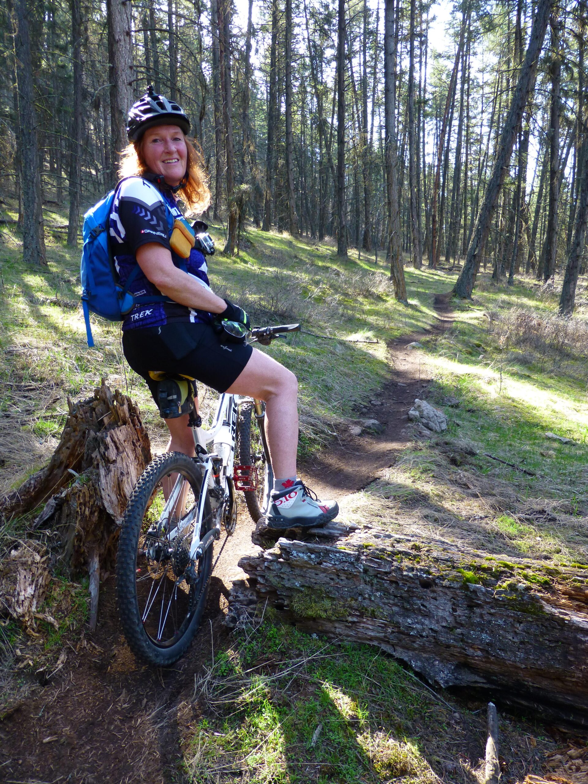 A person sitting on a mountain bike in a forested area, smiling at the camera. They are wearing a helmet, cycling gear, and a backpack. The trail is visible in the background, with tall trees and a sunny atmosphere. Ellison Park mountain bike trail.