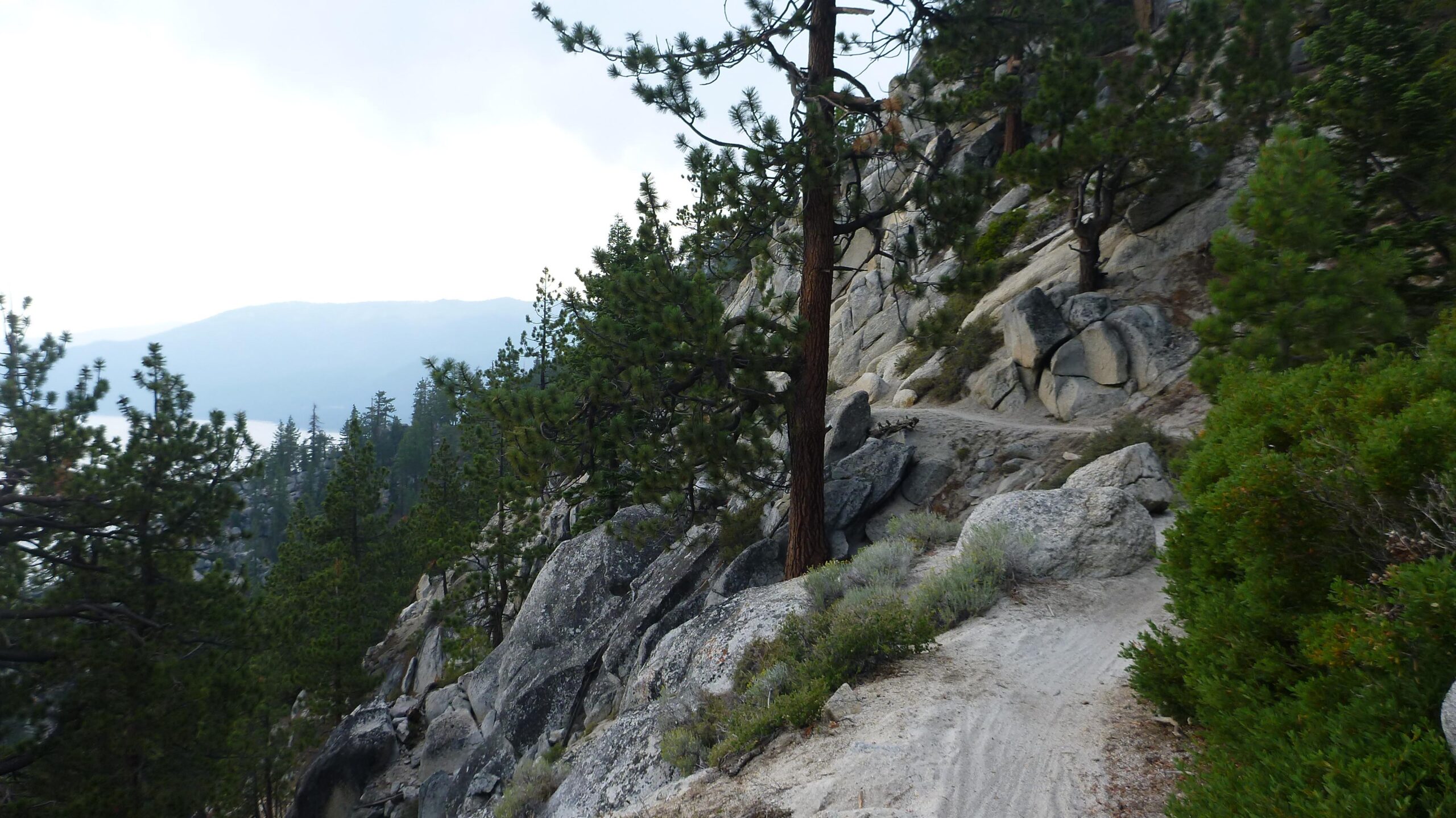 A winding dirt path bordered by rocks and pine trees, set against a backdrop of mountains and a cloudy sky. The scene captures the natural beauty of a rugged landscape. Tahoe Rim Trail: Tahoe Meadows to Tunnel Creek Road / Flume Trail mountain bike trail.