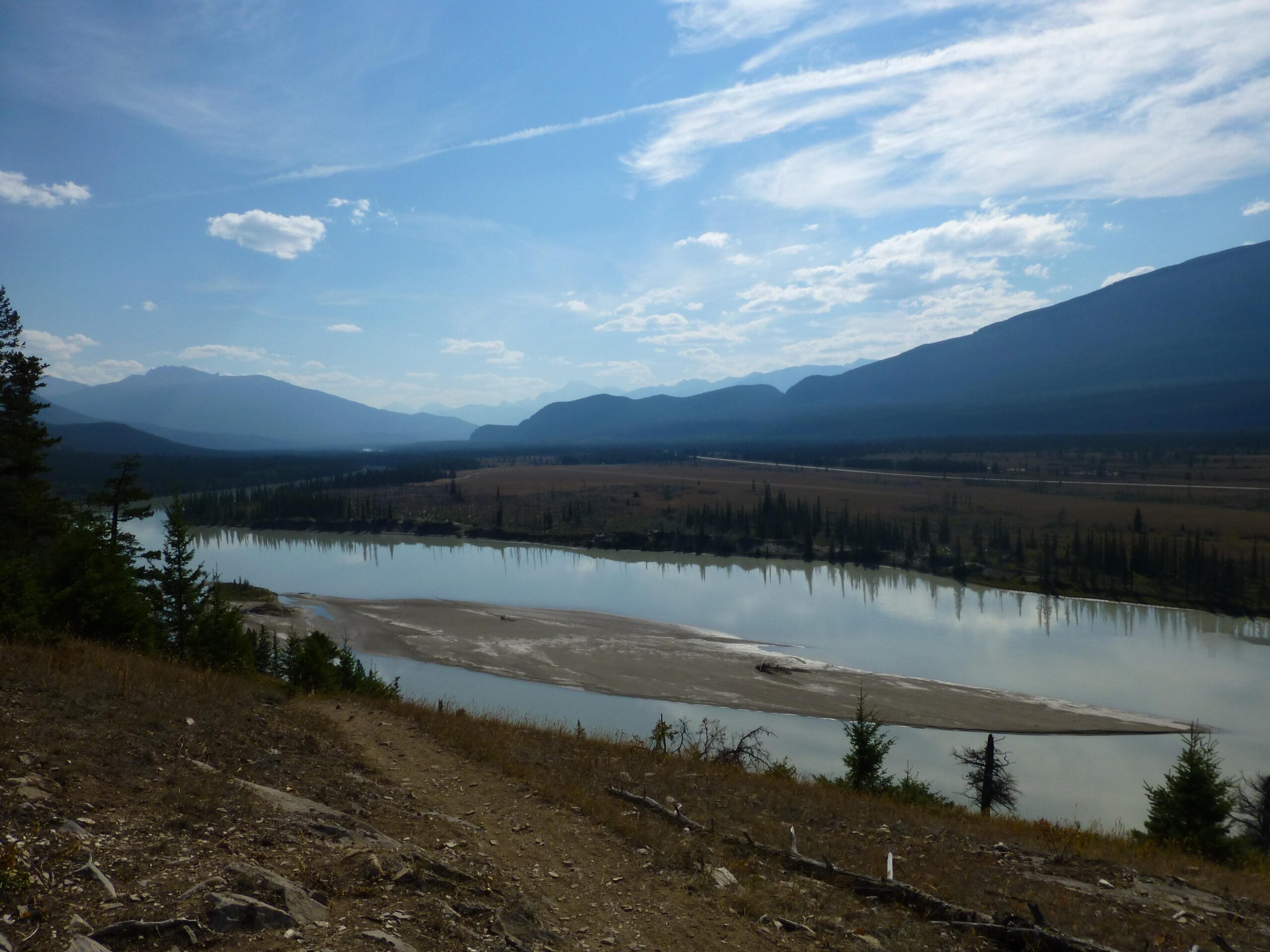 A scenic view of a river winding through a valley, surrounded by mountains under a partly cloudy sky. The calm water reflects the landscape, while trees and grassy banks frame the foreground. The Overlander mountain bike trail.