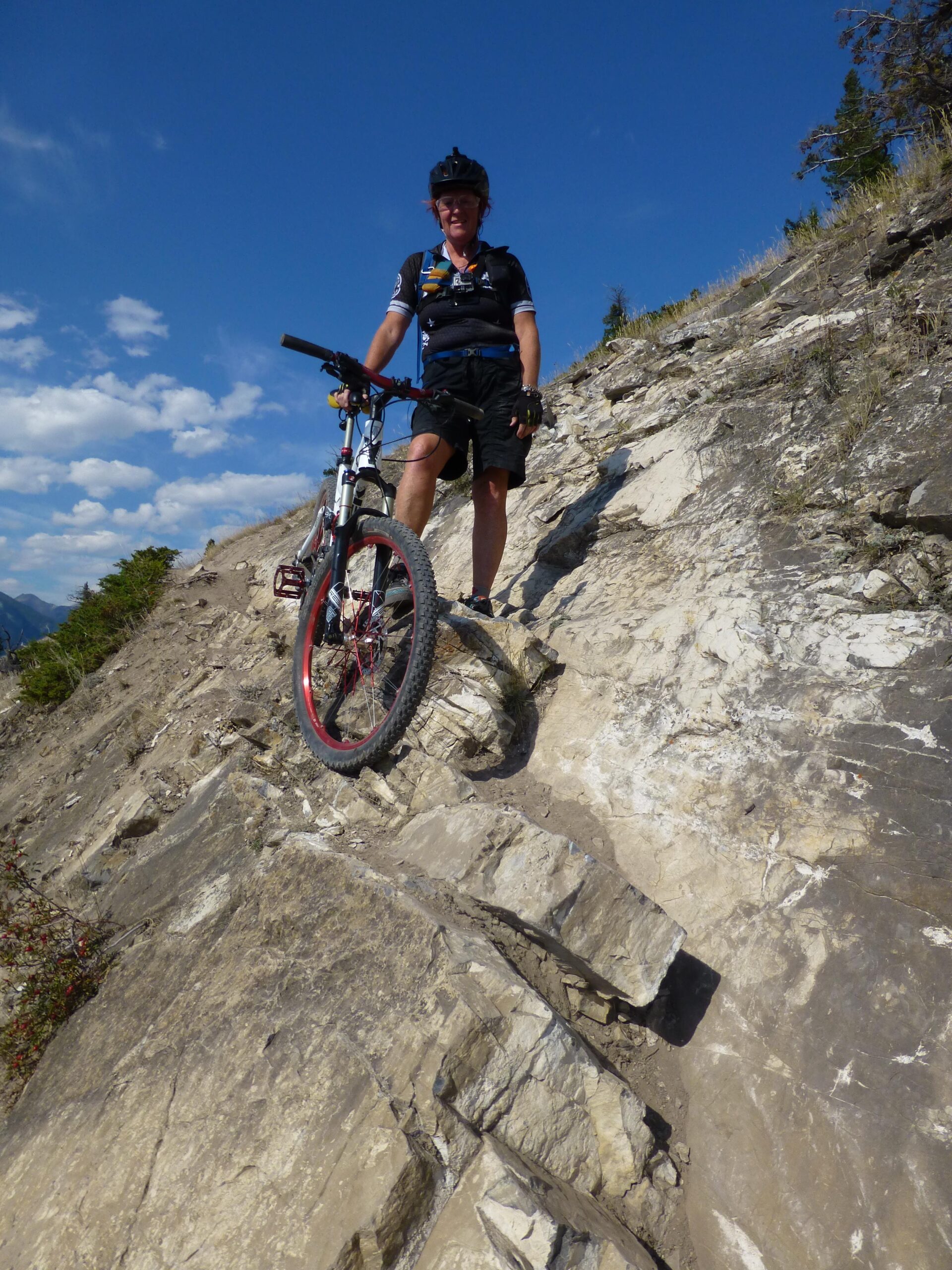 A mountain biker stands on a rocky trail, holding their bike while wearing a helmet and cycling gear. The landscape features steep, rocky terrain under a clear blue sky, with sparse vegetation in the background. The Overlander mountain bike trail.