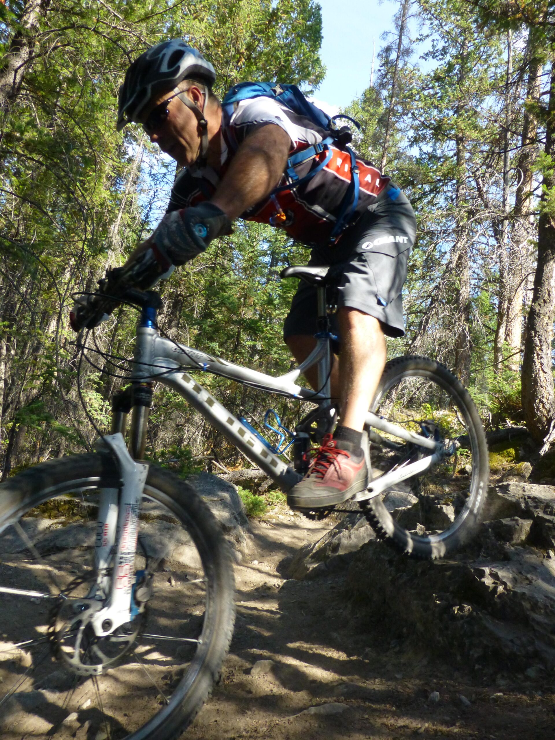 A male mountain biker in a helmet and protective gear rides over a rocky trail surrounded by trees. He leans forward as he navigates the terrain, showcasing the action and excitement of off-road cycling. The Overlander mountain bike trail.