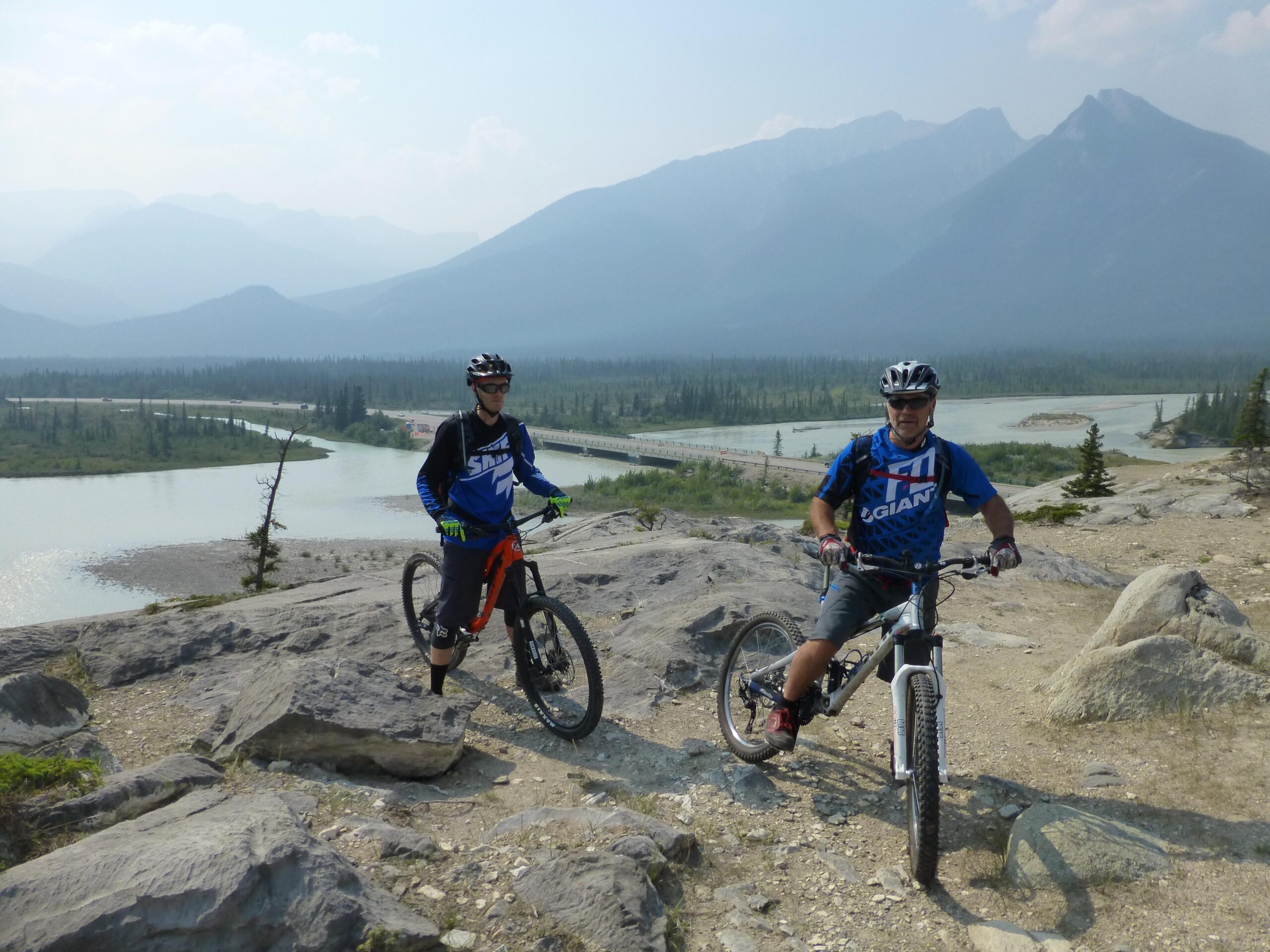Two mountain bikers stand on rocky terrain overlooking a winding river and lush green forest, with mountains in the background. One cyclist is wearing a blue and black jersey with sunglasses, while the other wears a blue and white jersey. The scene is set against a hazy sky, suggesting a warm day in a natural outdoor setting. The Overlander mountain bike trail.