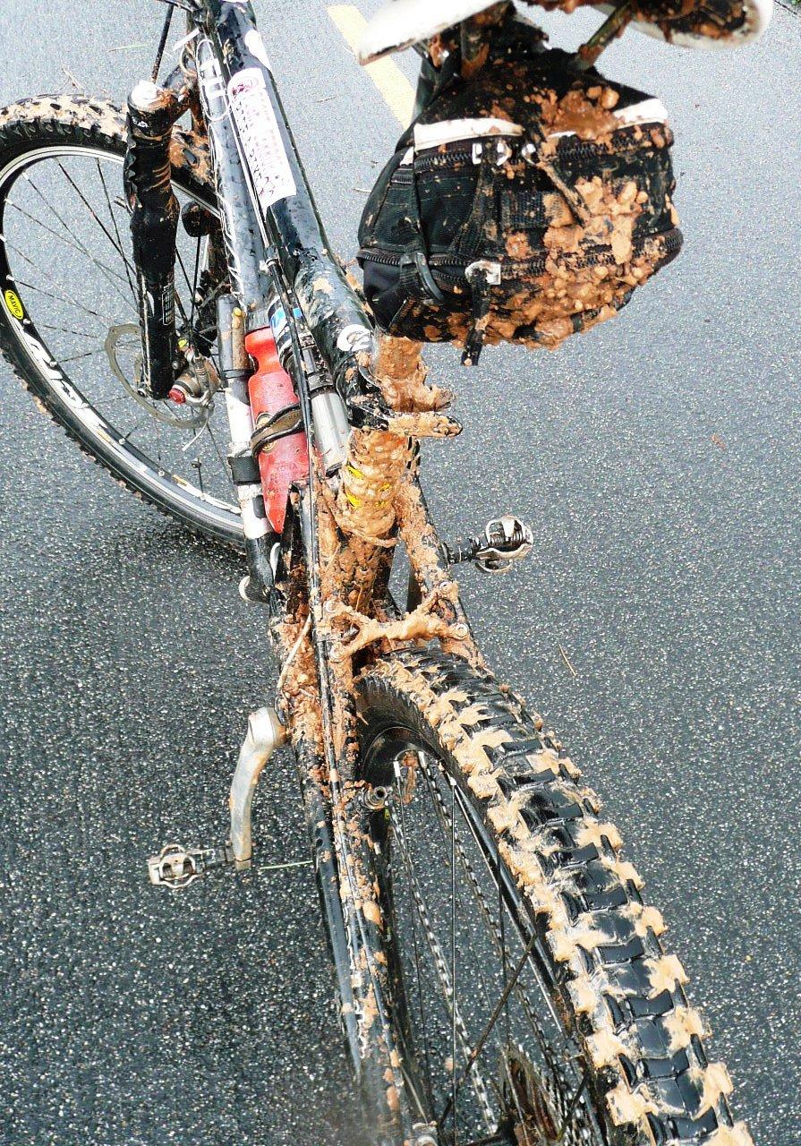 Santa Cruz Superlight: A close-up view of a muddy mountain bike, showcasing the rear section covered in dirt and mud. The bike features a visible water bottle, a black rear bag, and thick, knobby tires depicting heavy use on a wet, muddy trail. The background shows a wet road surface.