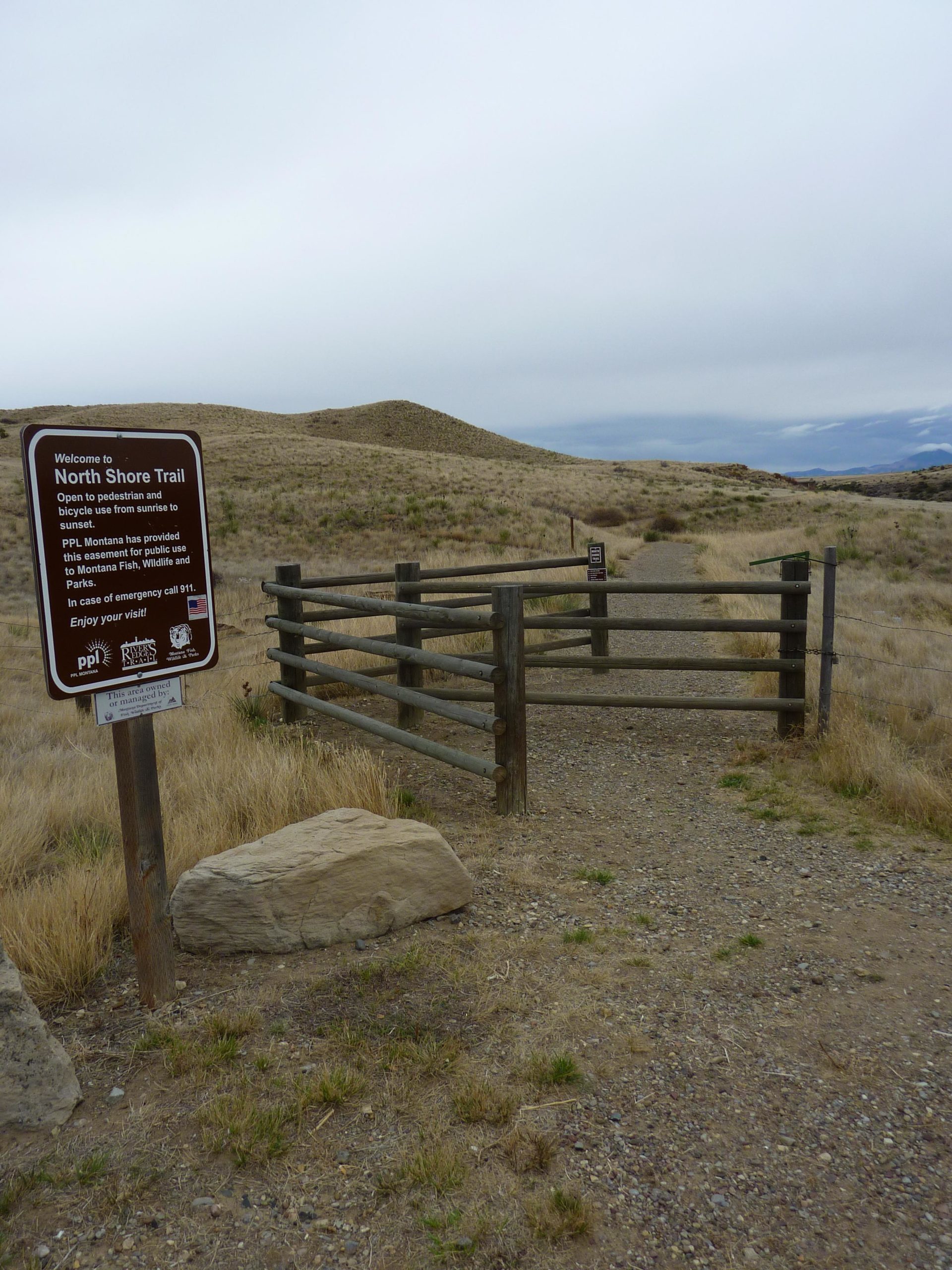 A dirt pathway leading into a natural area, marked by a wooden entrance gate. A large sign at the entrance reads "Welcome to North Shore Trail," indicating that the trail is open for pedestrian and bicycle use from sunrise to sunset. Surrounding the pathway are grassy hills under a cloudy sky. North Shore mountain bike trail.