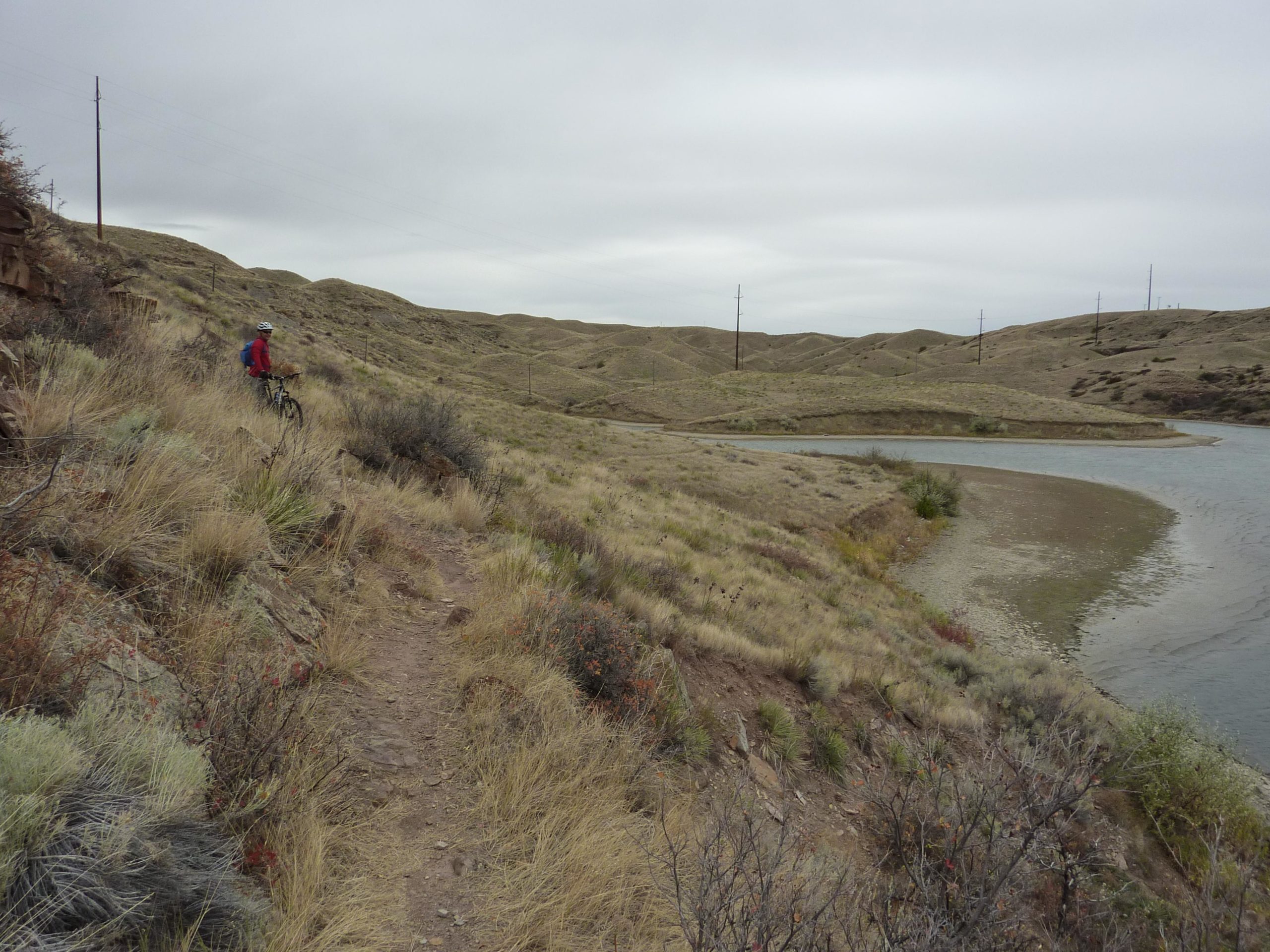 A cyclist in a red jacket riding along a narrow trail beside a river, surrounded by rolling hills and sparse vegetation under a cloudy sky. Electric poles are visible in the background, contributing to the remote landscape. North Shore mountain bike trail.