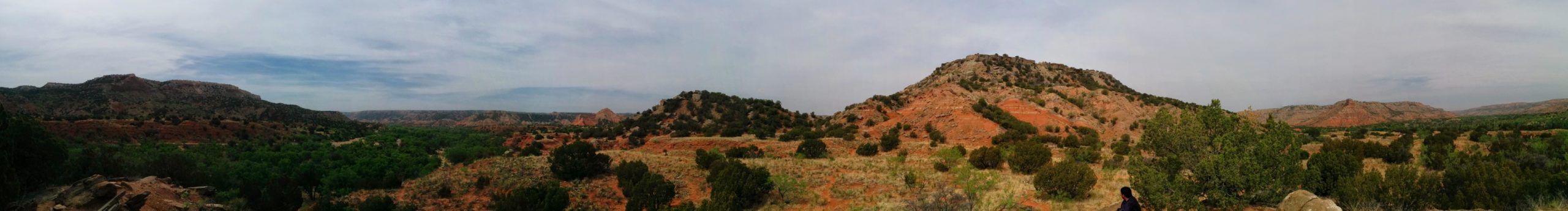 Panoramic view of a colorful landscape featuring rolling hills and rock formations, with greenery in the foreground. The sky is partly cloudy, showcasing a serene and natural setting. Palo Duro Canyon mountain bike trail.