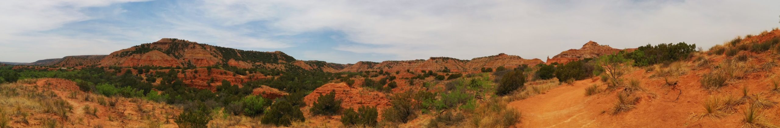 A panoramic view of a rugged landscape featuring reddish-brown rock formations and sparse greenery under a partially cloudy sky. The scene captures the natural beauty of the terrain with rolling hills, bushes, and a dirt path winding through the foreground. Palo Duro Canyon mountain bike trail.